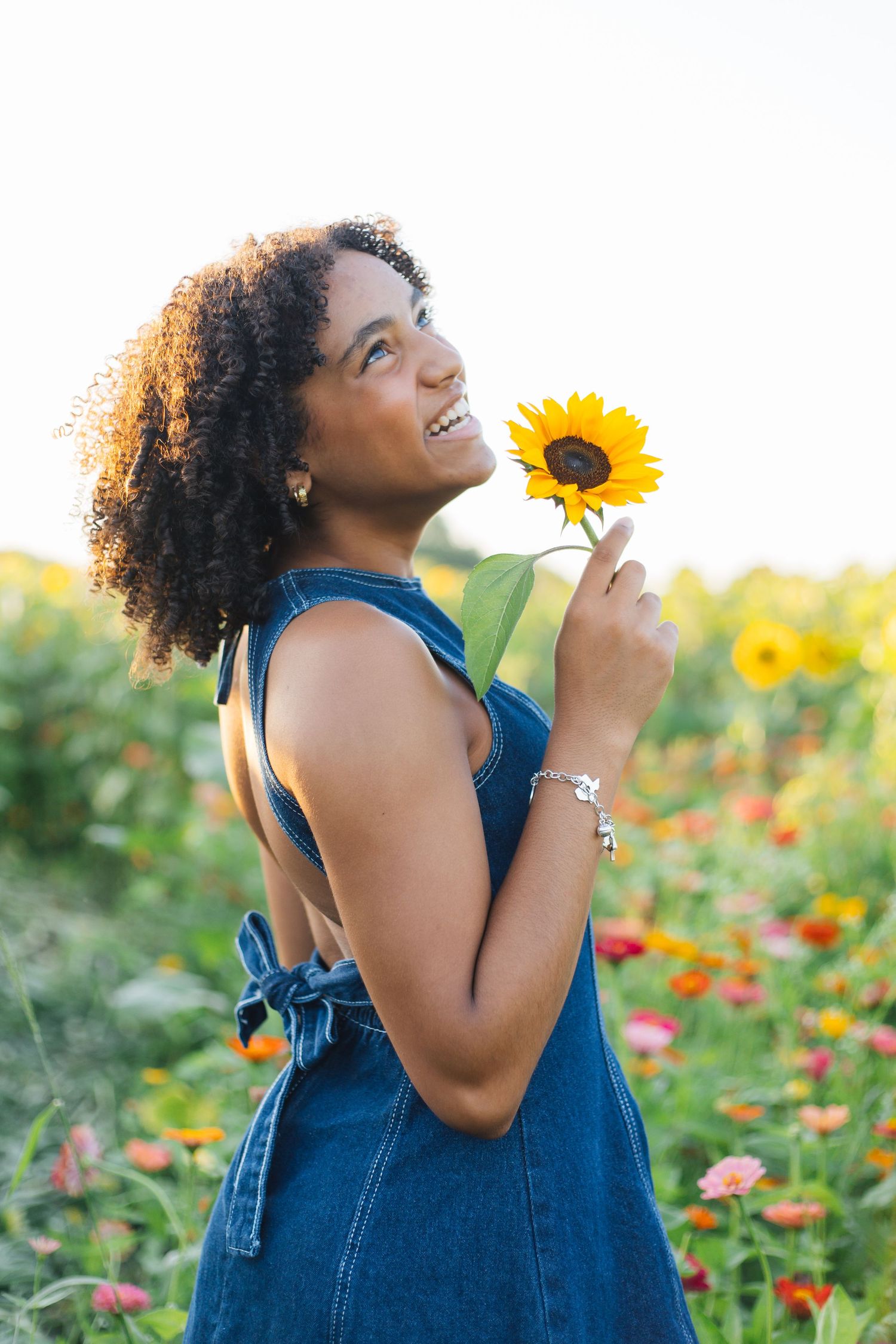 Smiling woman in denim dress holding a sunflower in a colorful flower field on a sunny day.