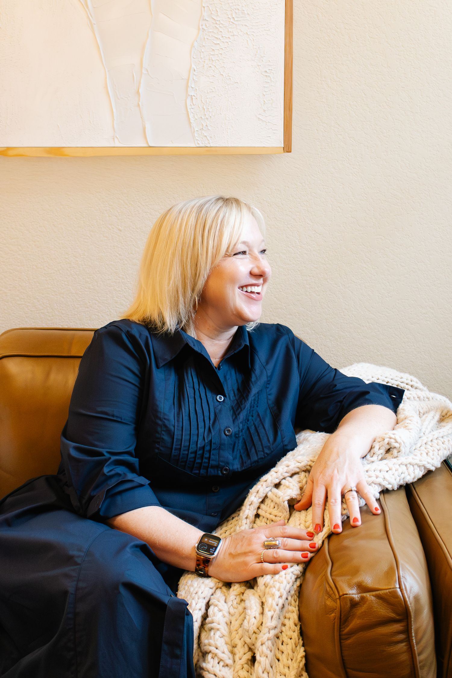 Smiling blonde woman in navy dress sitting on brown leather sofa with chunky knit blanket, laughing in cozy interior.