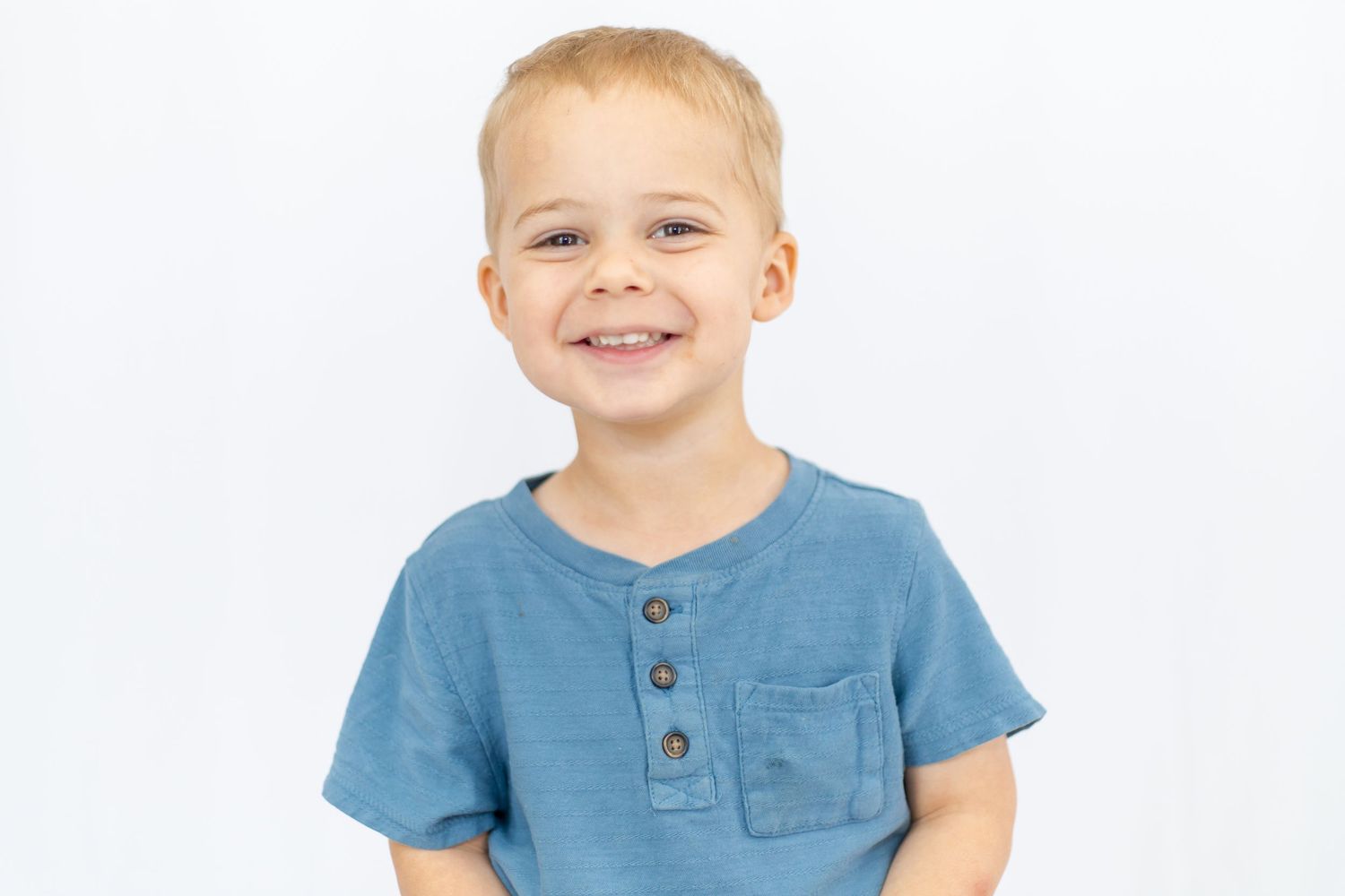 Smiling young boy with blonde hair wearing a blue henley t-shirt against a white background.