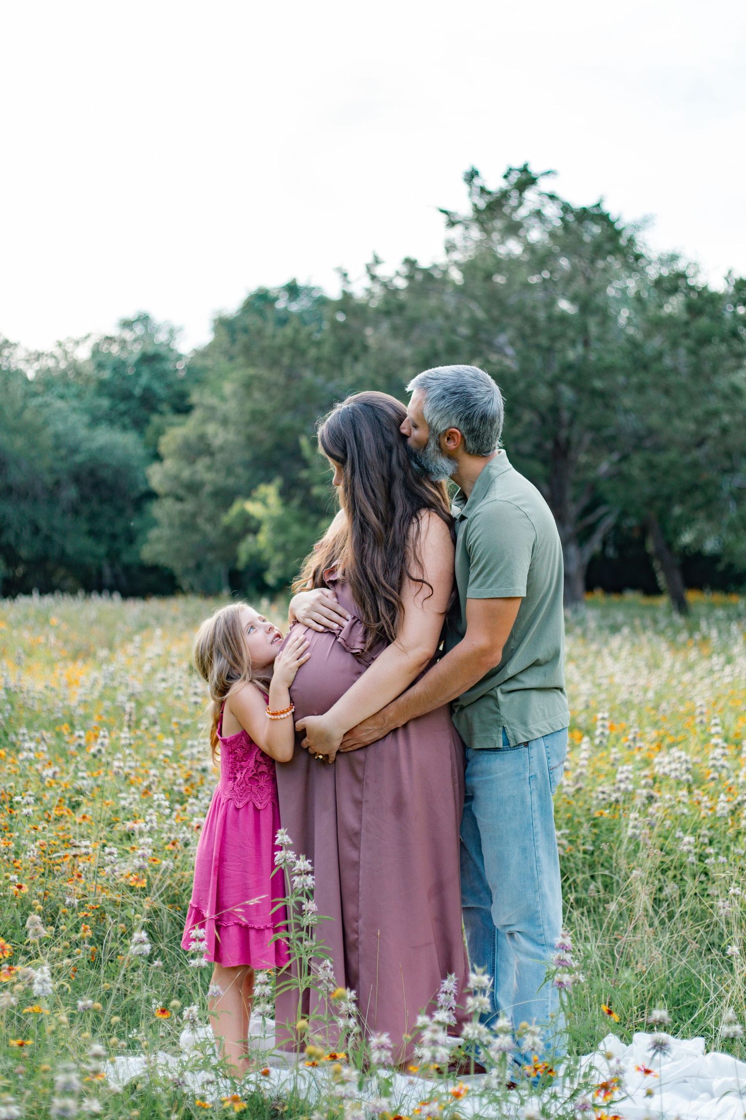 A family maternity photo in a wildflower field, with a young girl hugging her pregnant mother's belly while father kisses her head.