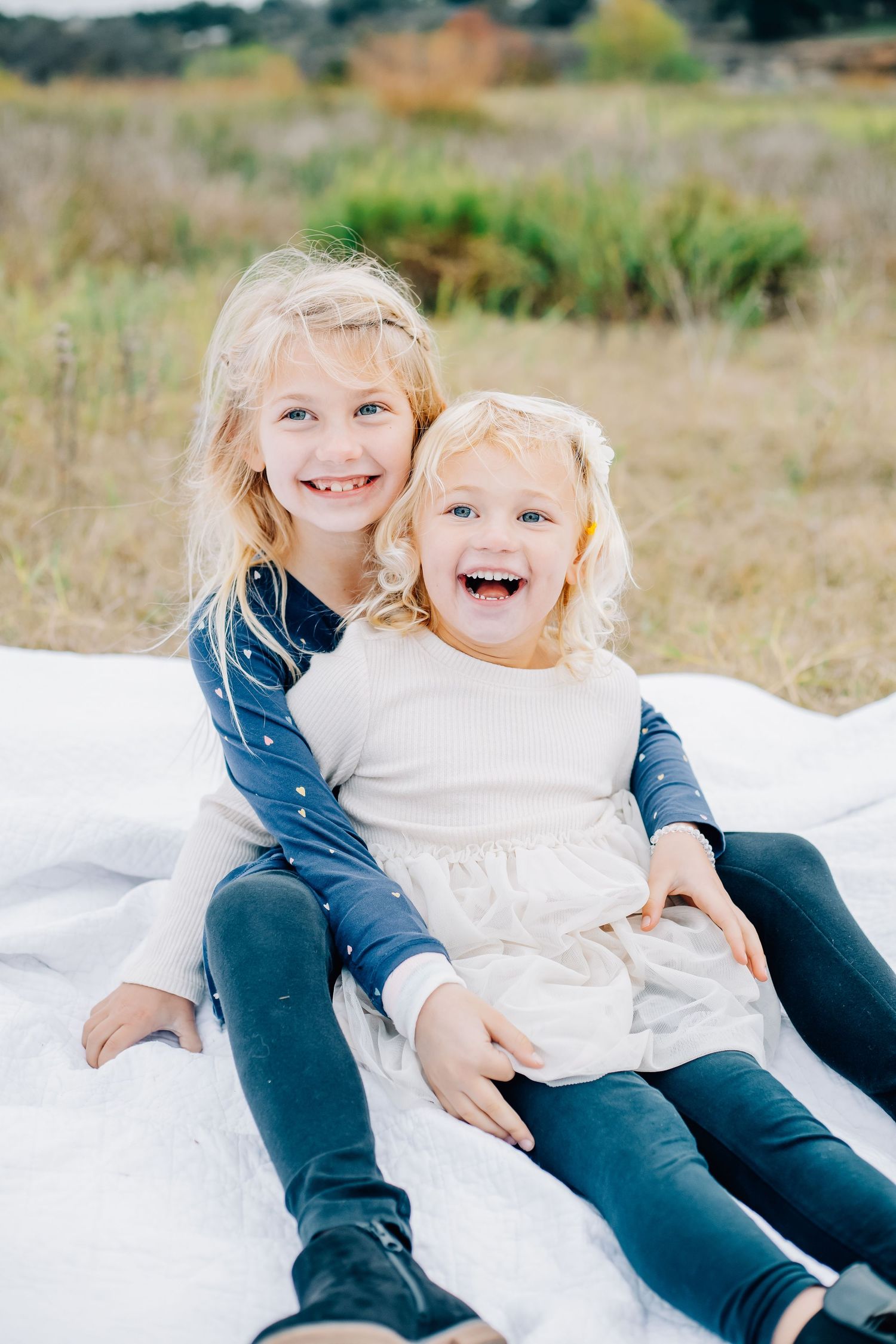 Two children laughing and playing together on a white blanket outdoors in a natural setting.