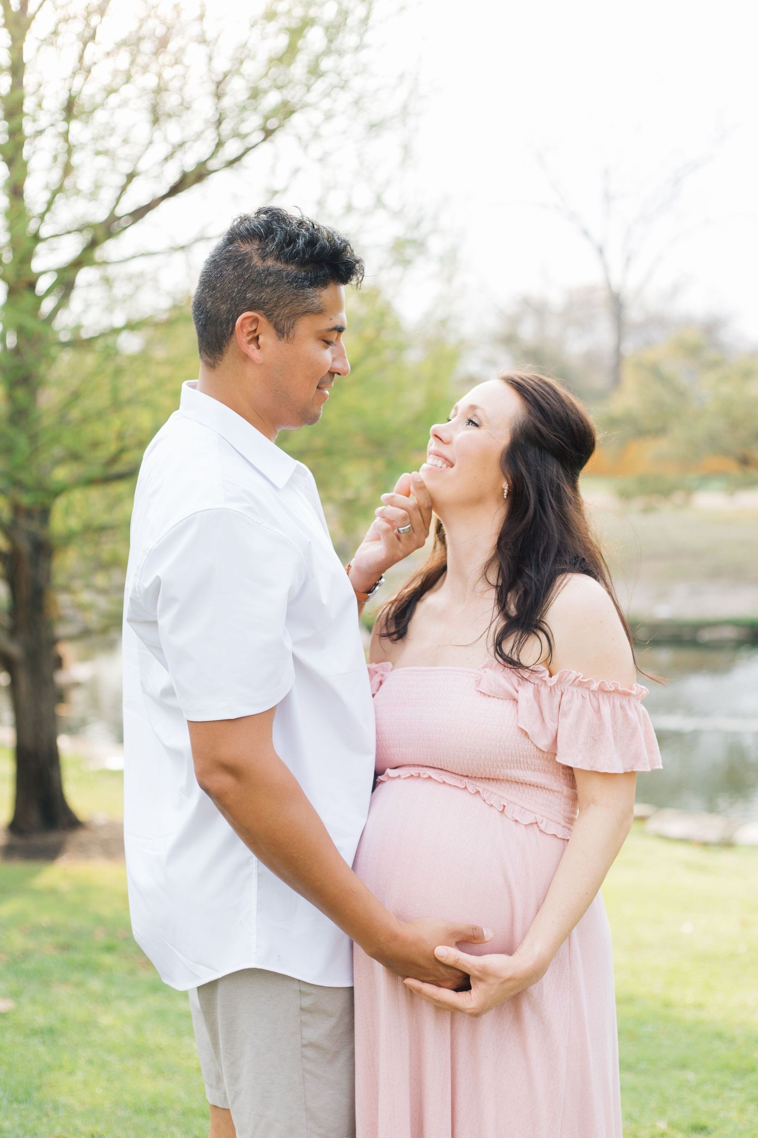 A couple embraces during a maternity photoshoot near a lake on a spring afternoon.
