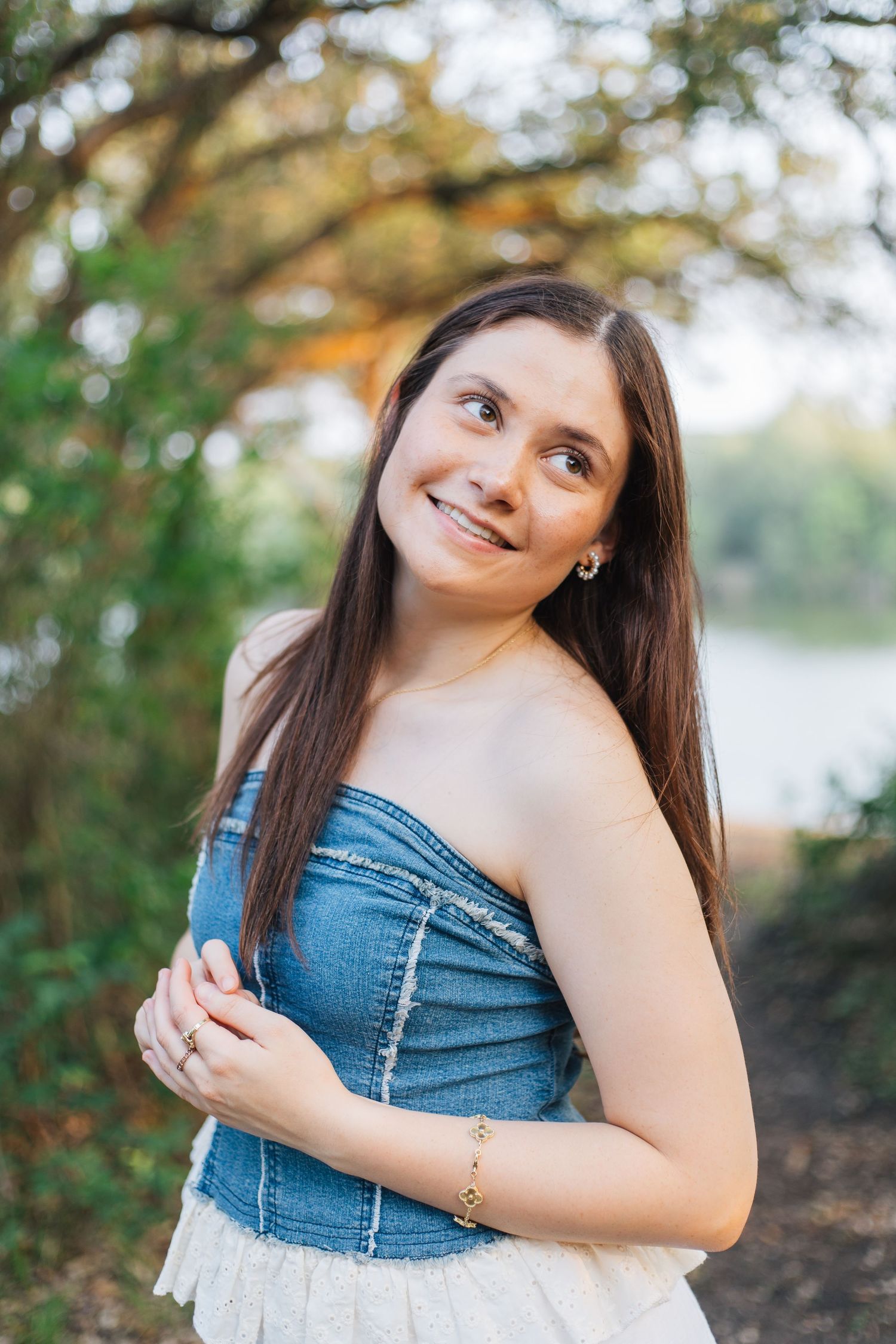 Young woman in denim strapless top smiling outdoors near trees and lake in natural setting. Senior Portraits Round Rock TX