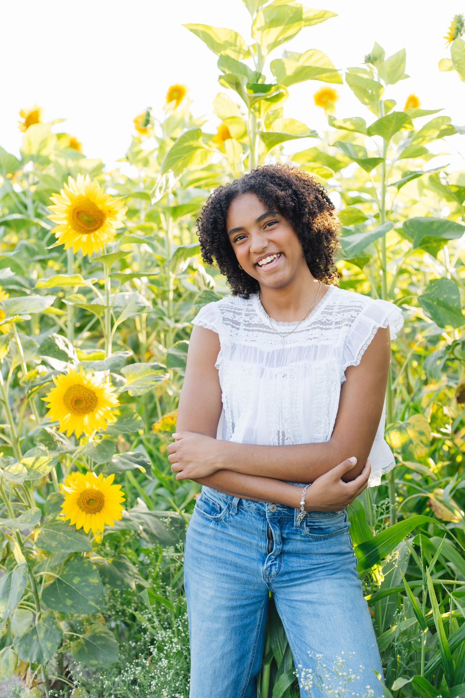 Smiling young woman with curly hair in white top and jeans posing with arms crossed in a sunflower field.