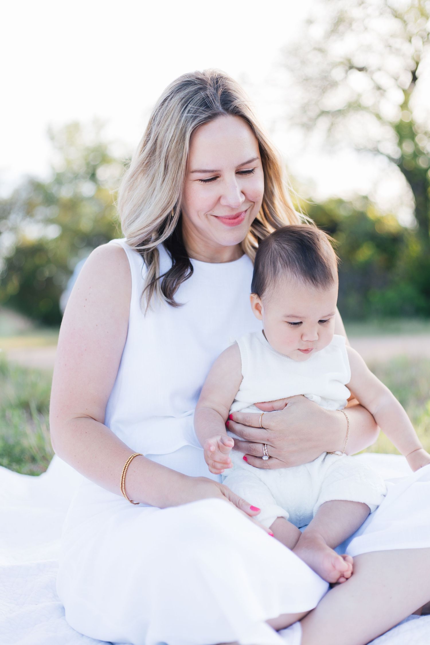 Smiling blonde mother in white dress sits outdoors cradling her infant baby in natural light.