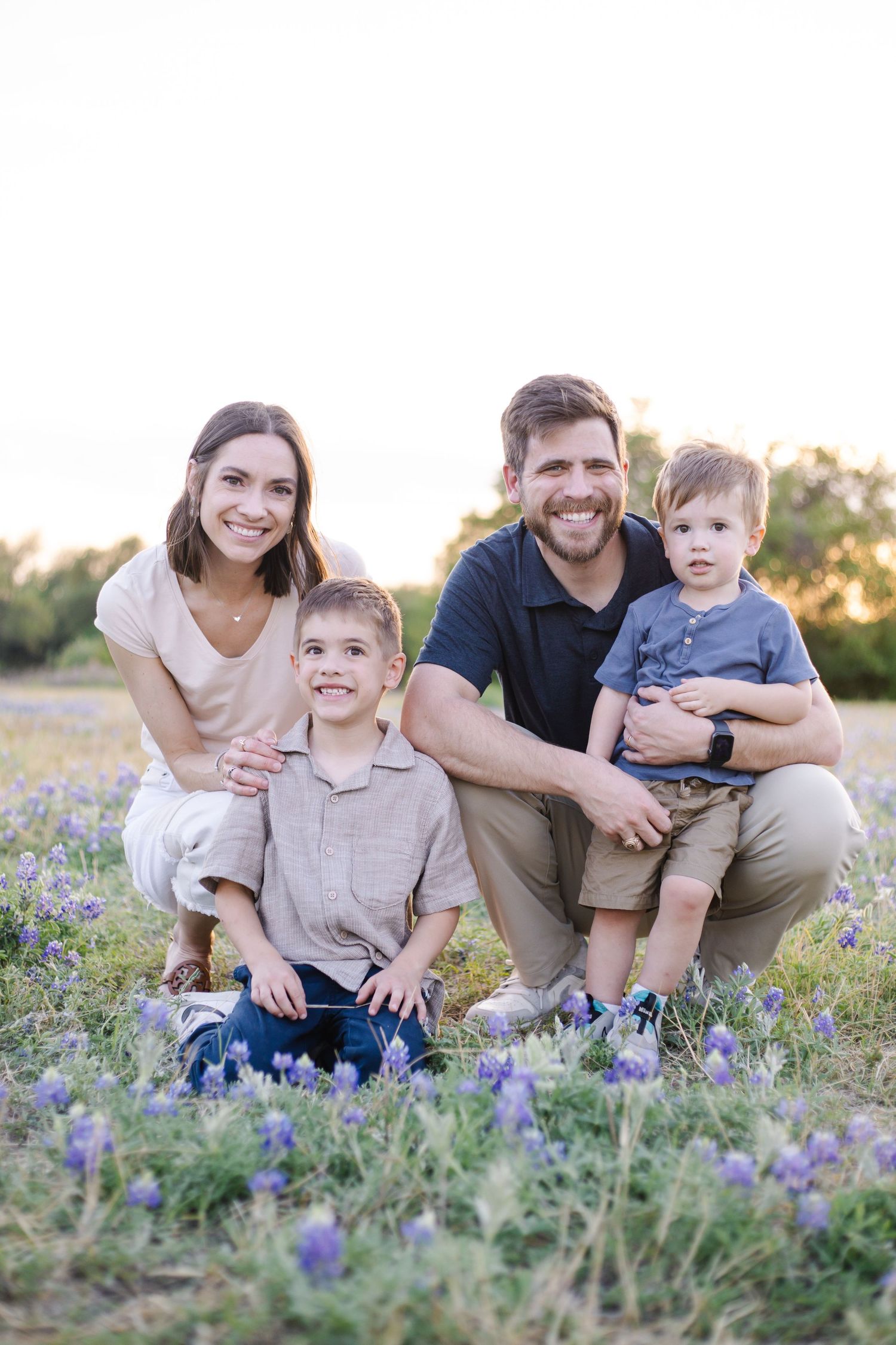 Smiling family of four crouches together in a field of purple wildflowers during golden hour sunset.