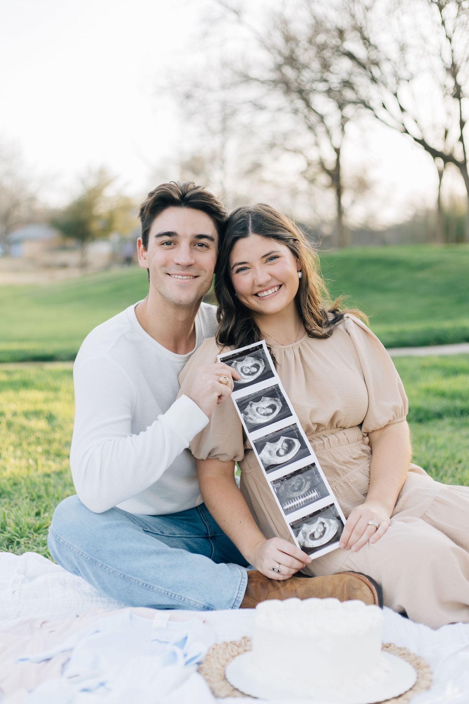 Couple sits on picnic blanket outdoors holding ultrasound strip showing pregnancy announcement.