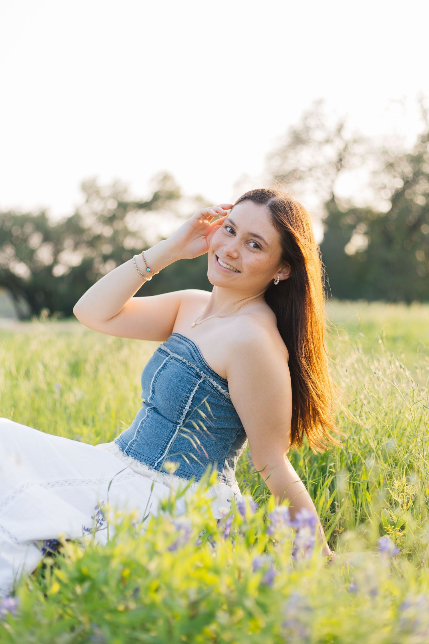 Young woman in denim corset top and white skirt sitting in green meadow with wildflowers, smiling in golden sunlight. Senior Portraits