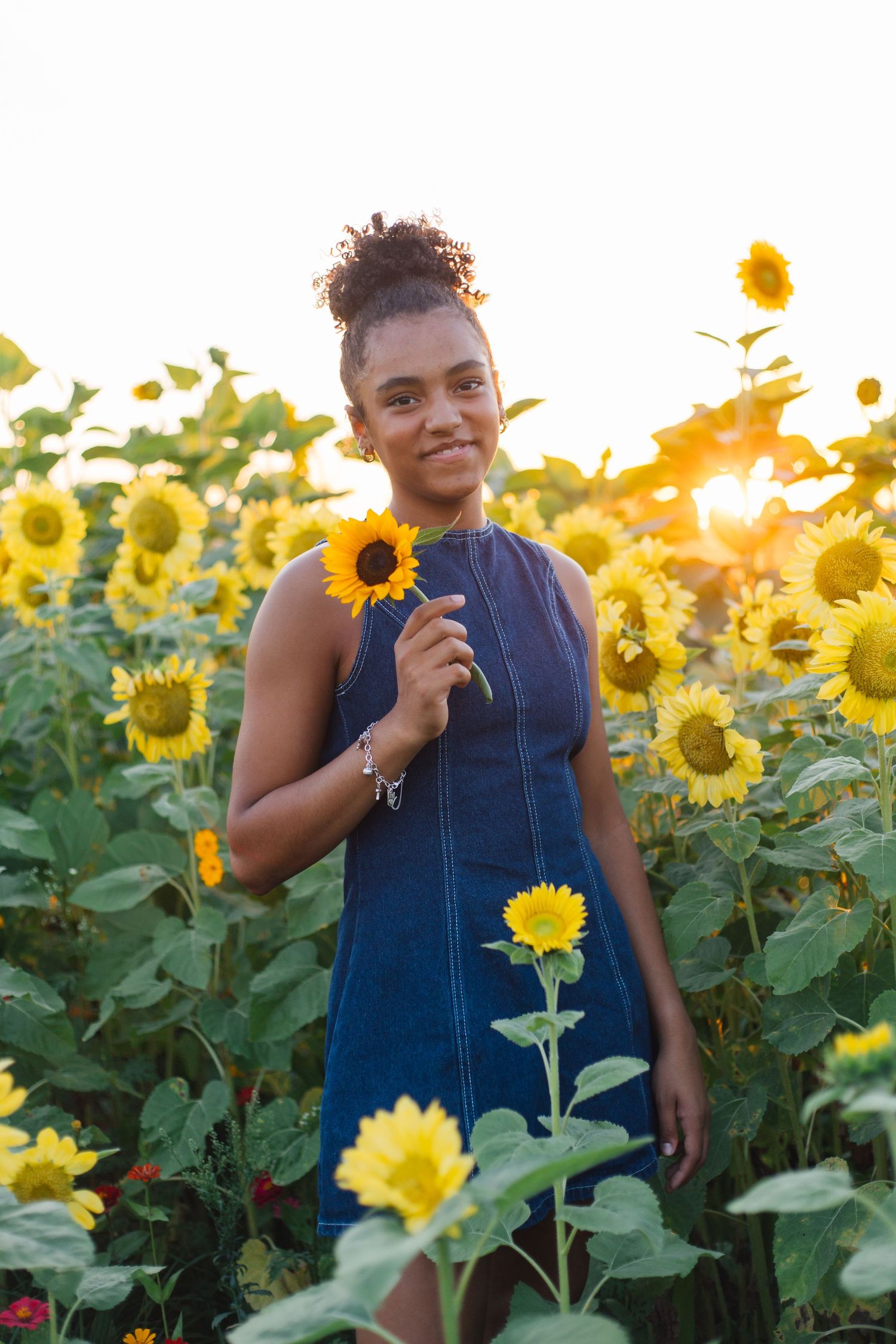 Young Black woman in denim dress holding sunflower in golden sunlit field, smiling warmly at camera.