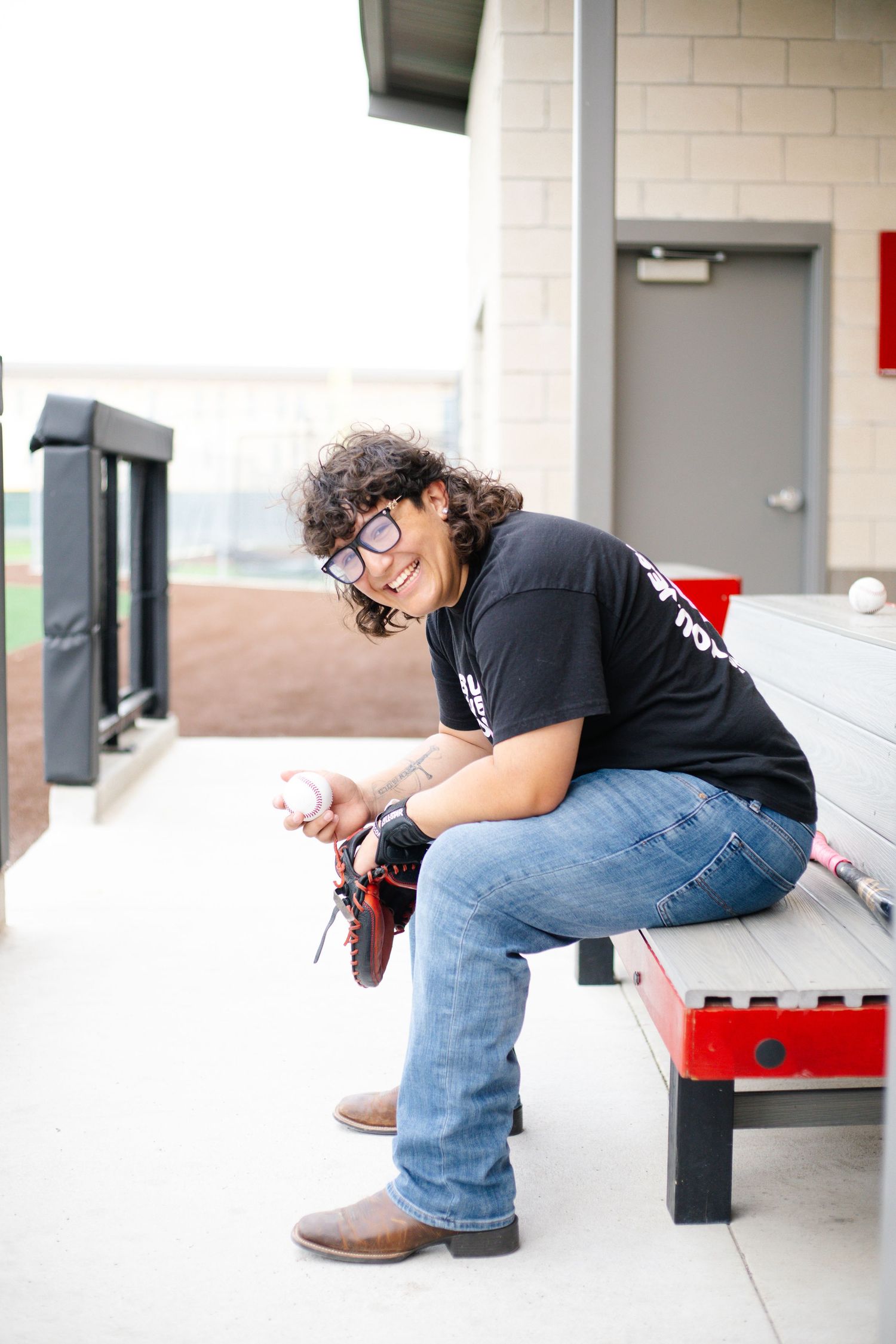 Young athlete sits on baseball dugout bench smiling while holding a baseball and glove in jeans and black tee.