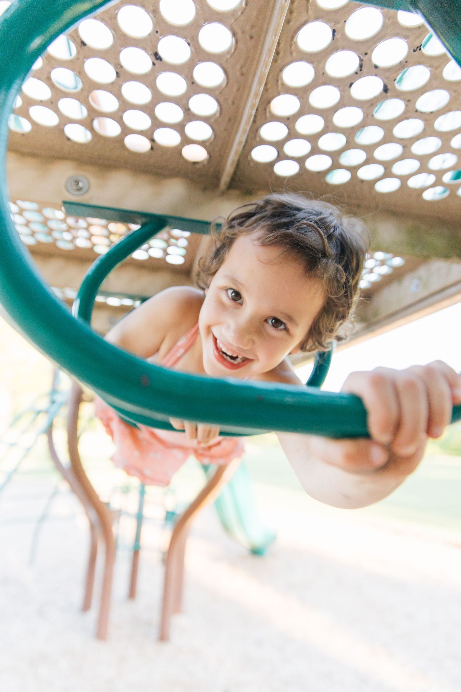 A happy young girl climbing playground equipment, smiling up at the camera while gripping a green metal railing.