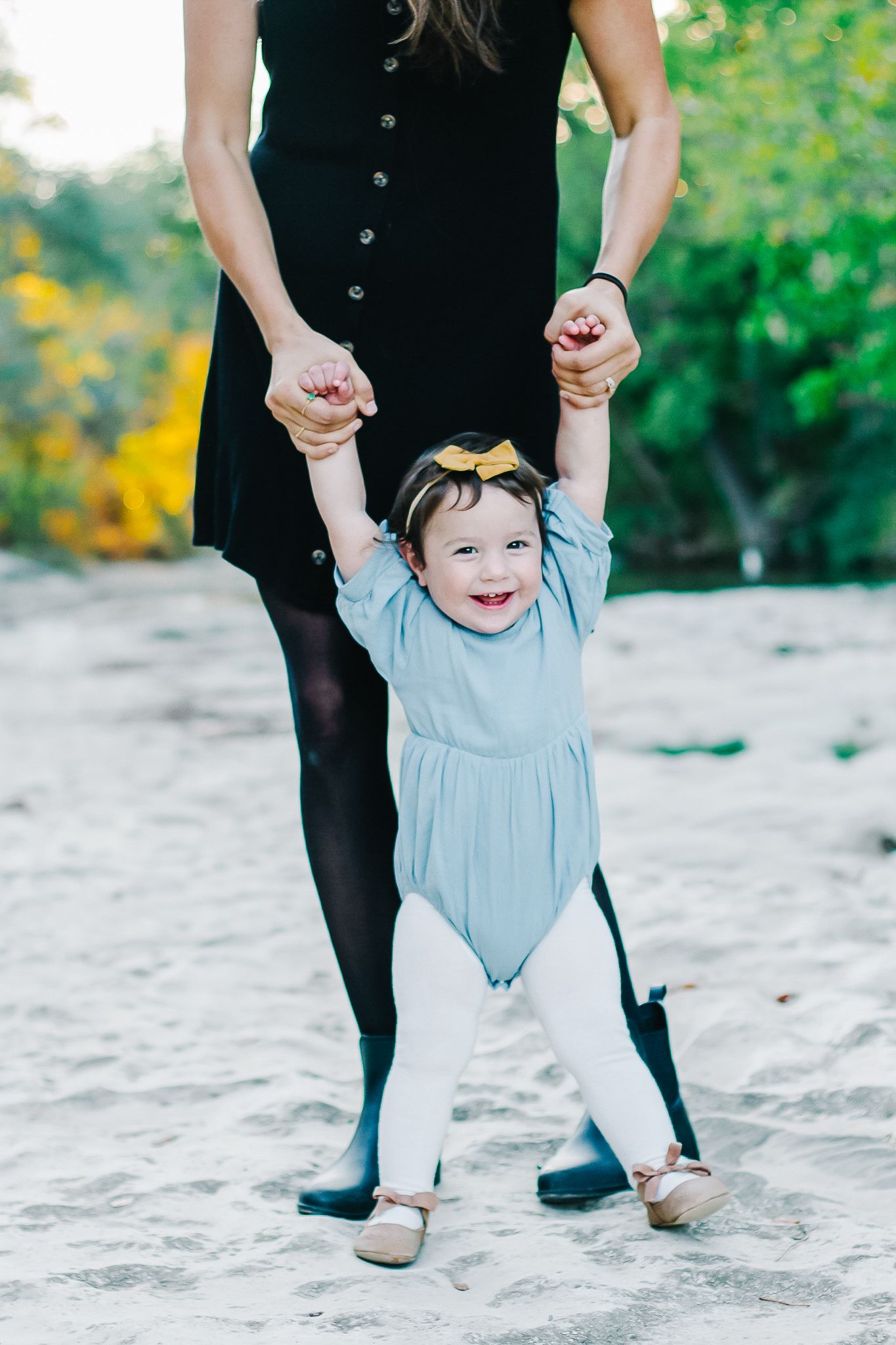 Small child in blue dress taking supported steps on beach while holding hands with adult.