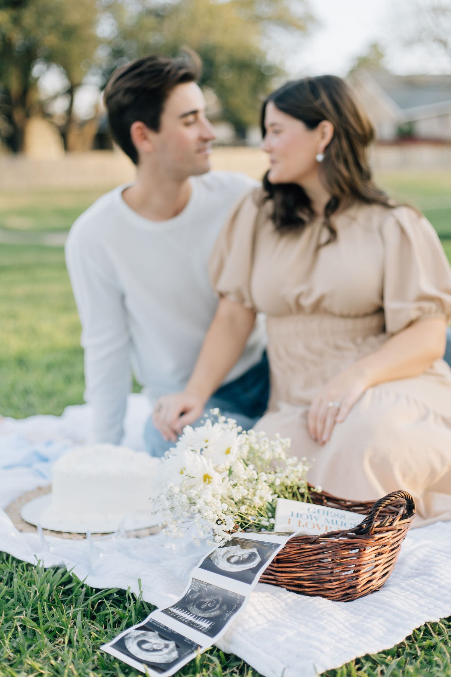 Couple enjoys romantic picnic on blanket with ultrasound photos and white flowers.