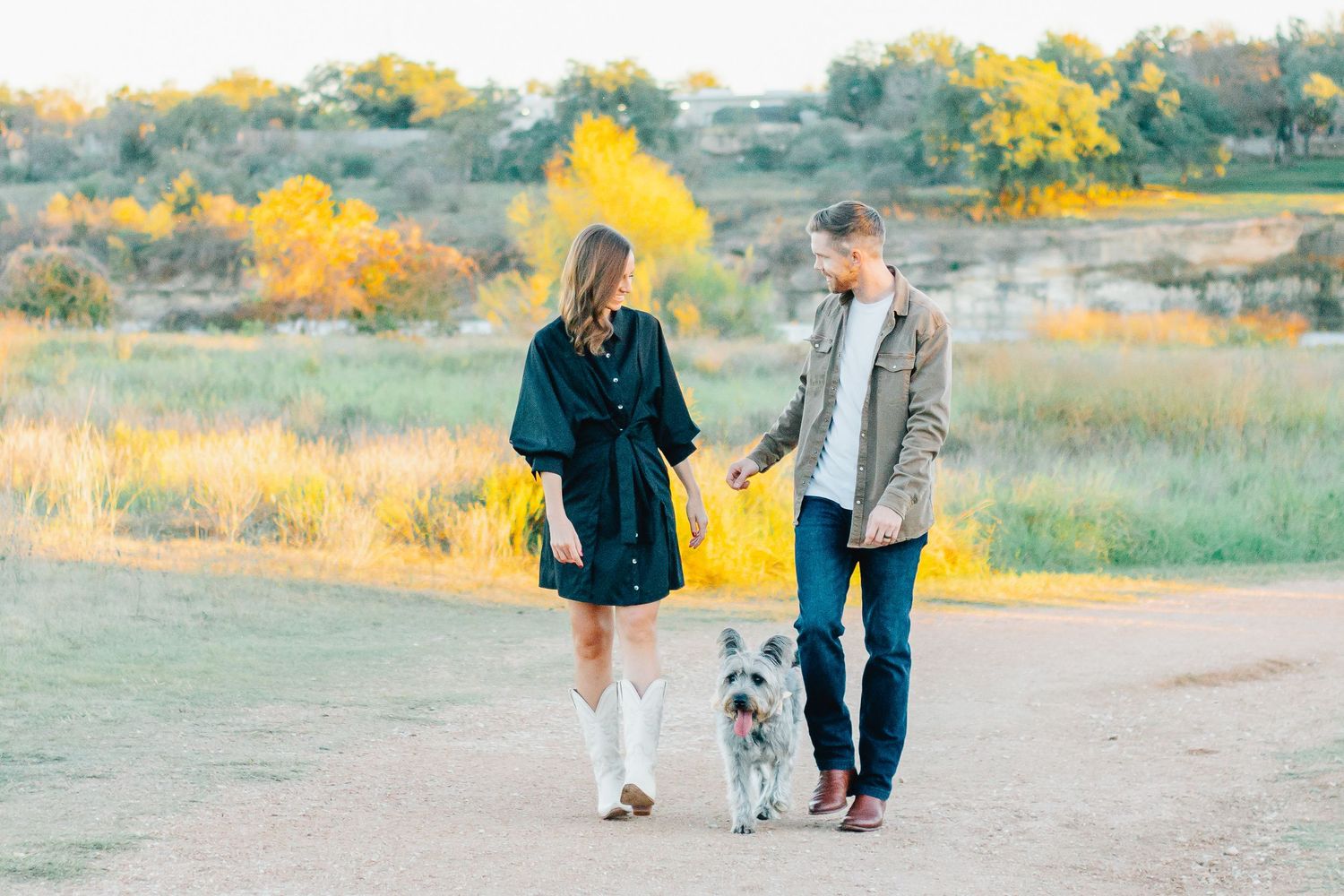 A couple and their dog walk together in a golden field during sunset.
