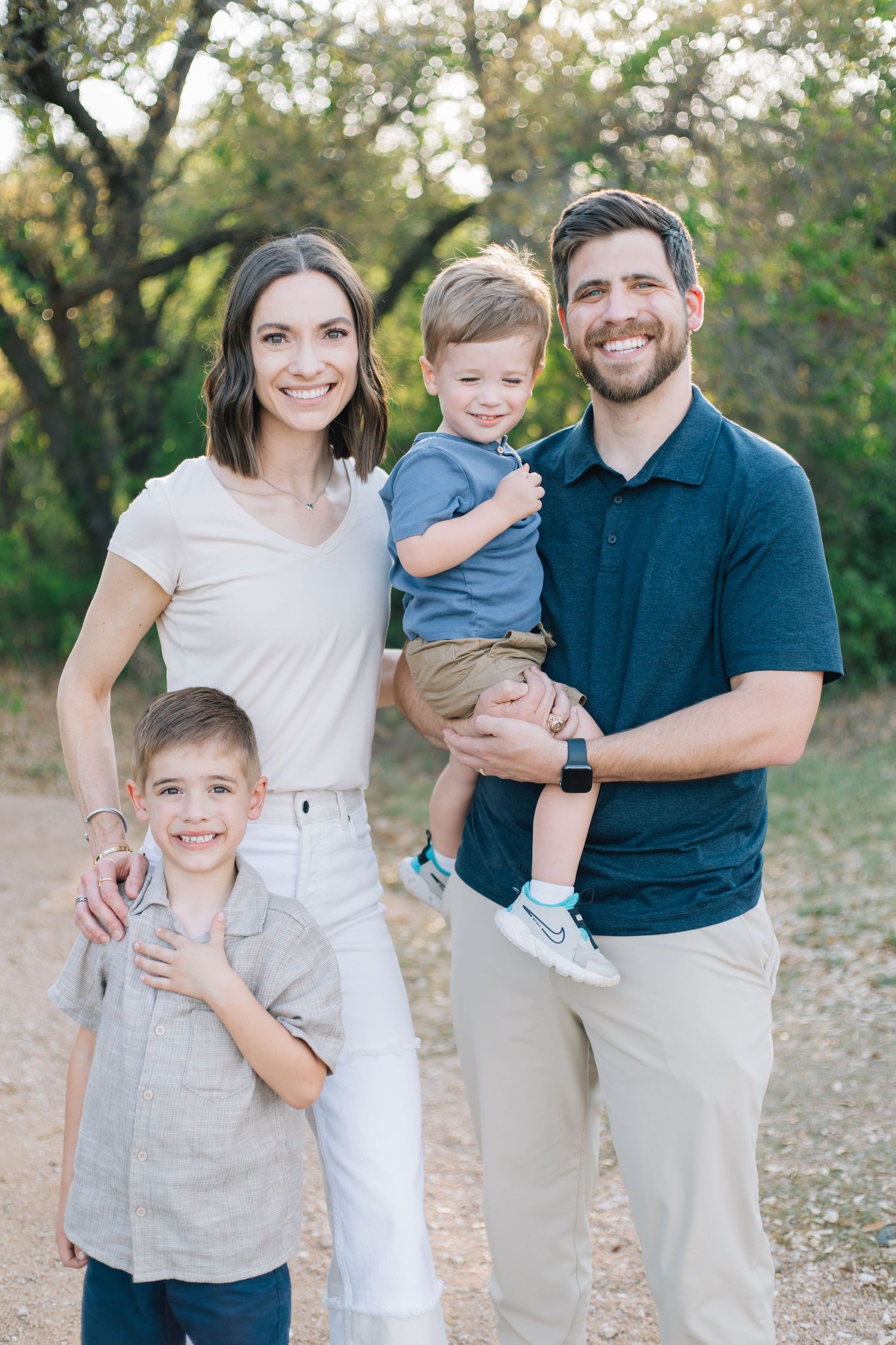 Happy family of four poses outdoors on a dirt path, parents smiling with two young boys among green trees.