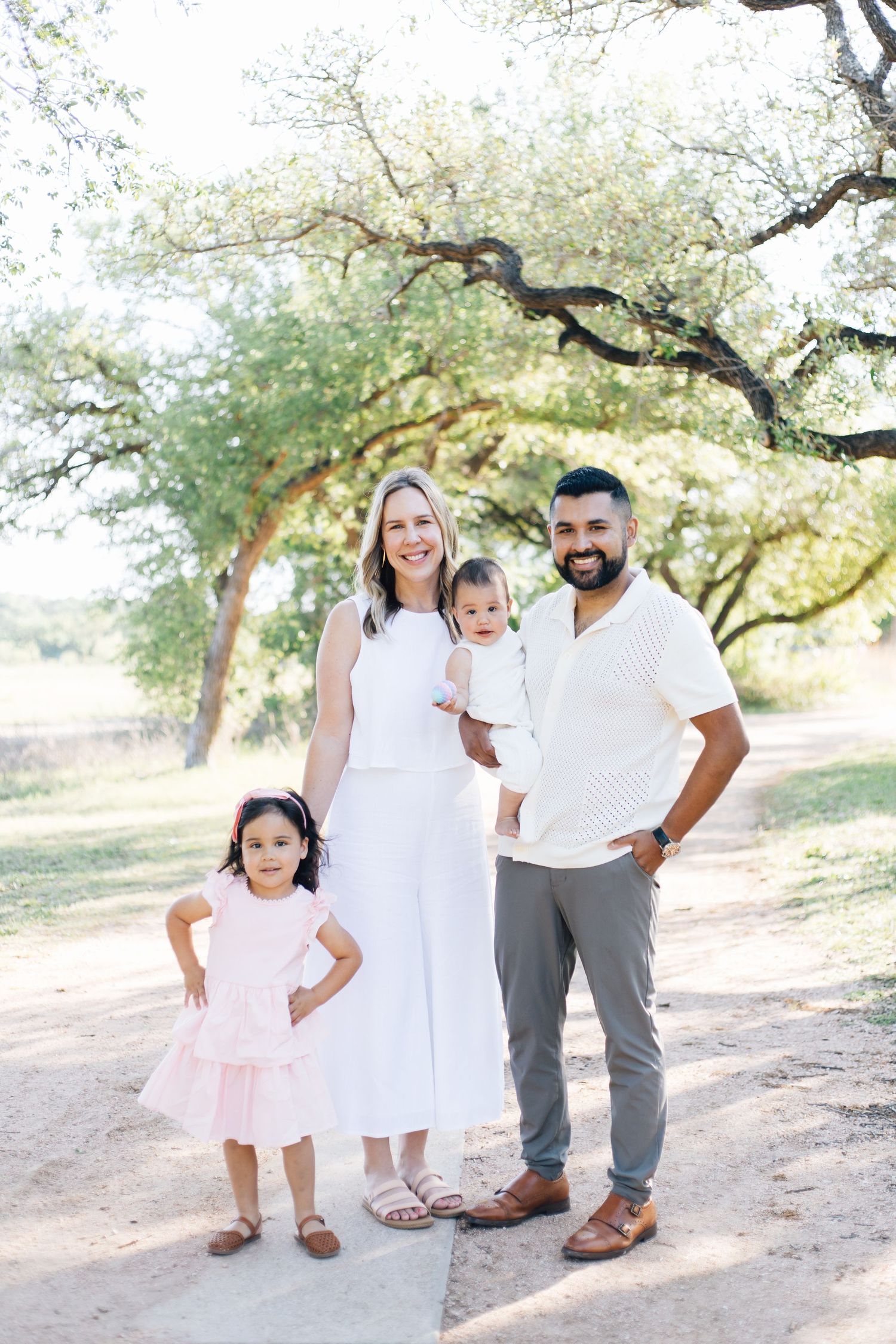 Family of four poses on a sunny tree-lined path, parents in white with two young children.