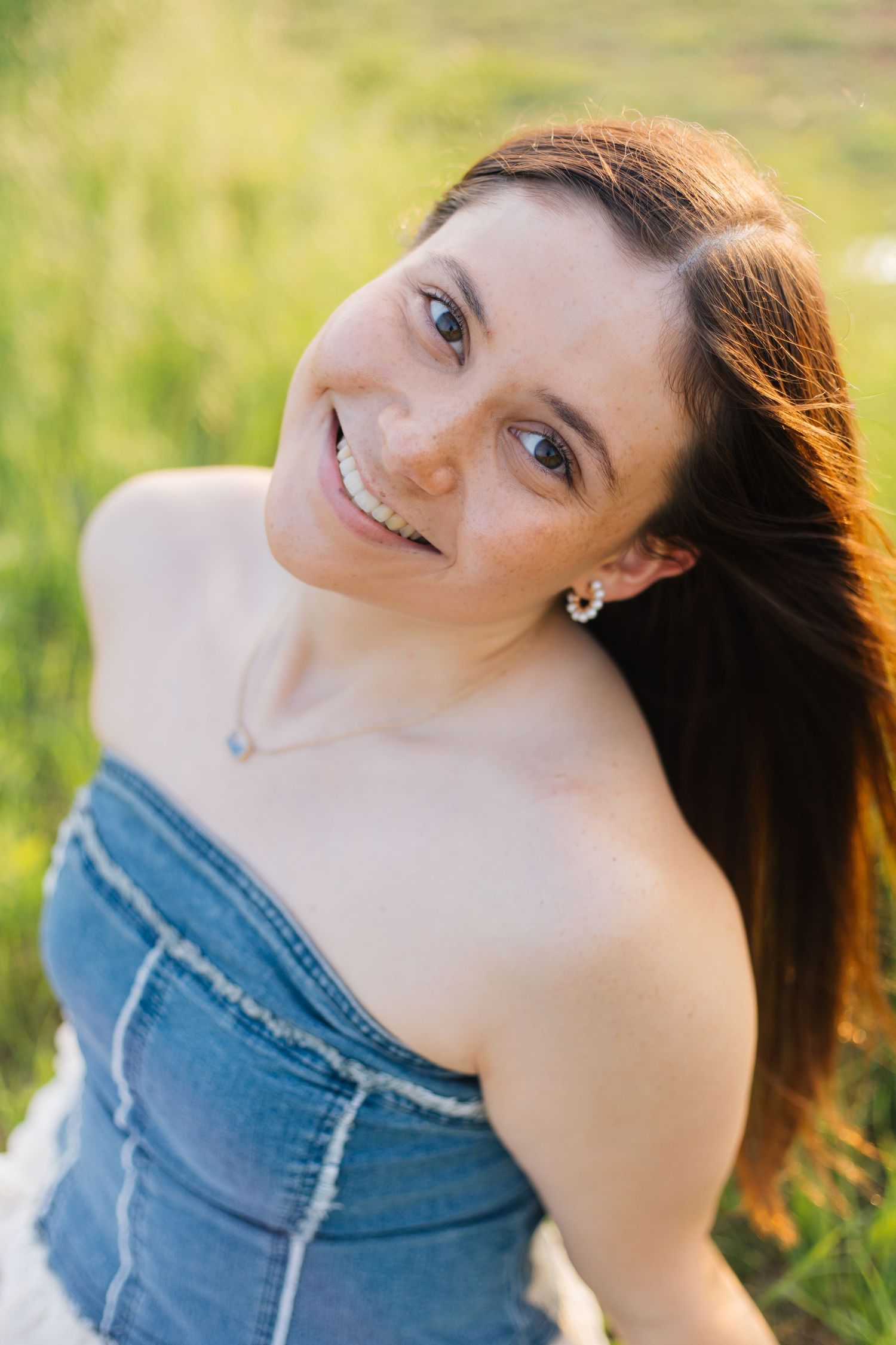 Smiling young woman with long brown hair wearing a denim tube top outdoors in a sunny green field. Senior Portraits Round Rock TX