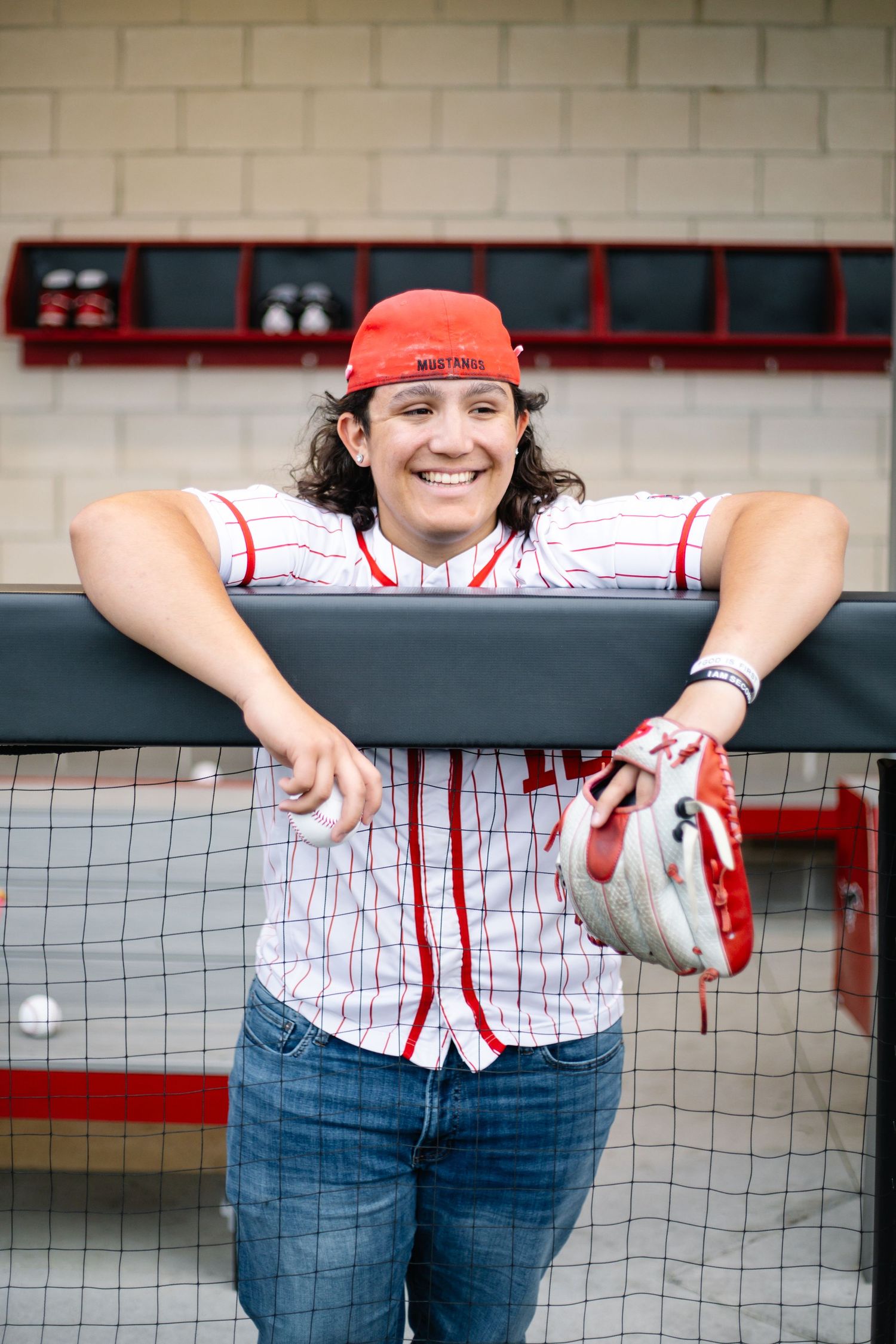 Smiling softball player in red cap and pinstripe jersey leans on dugout fence holding a glove. Senior Portraits Austin TX