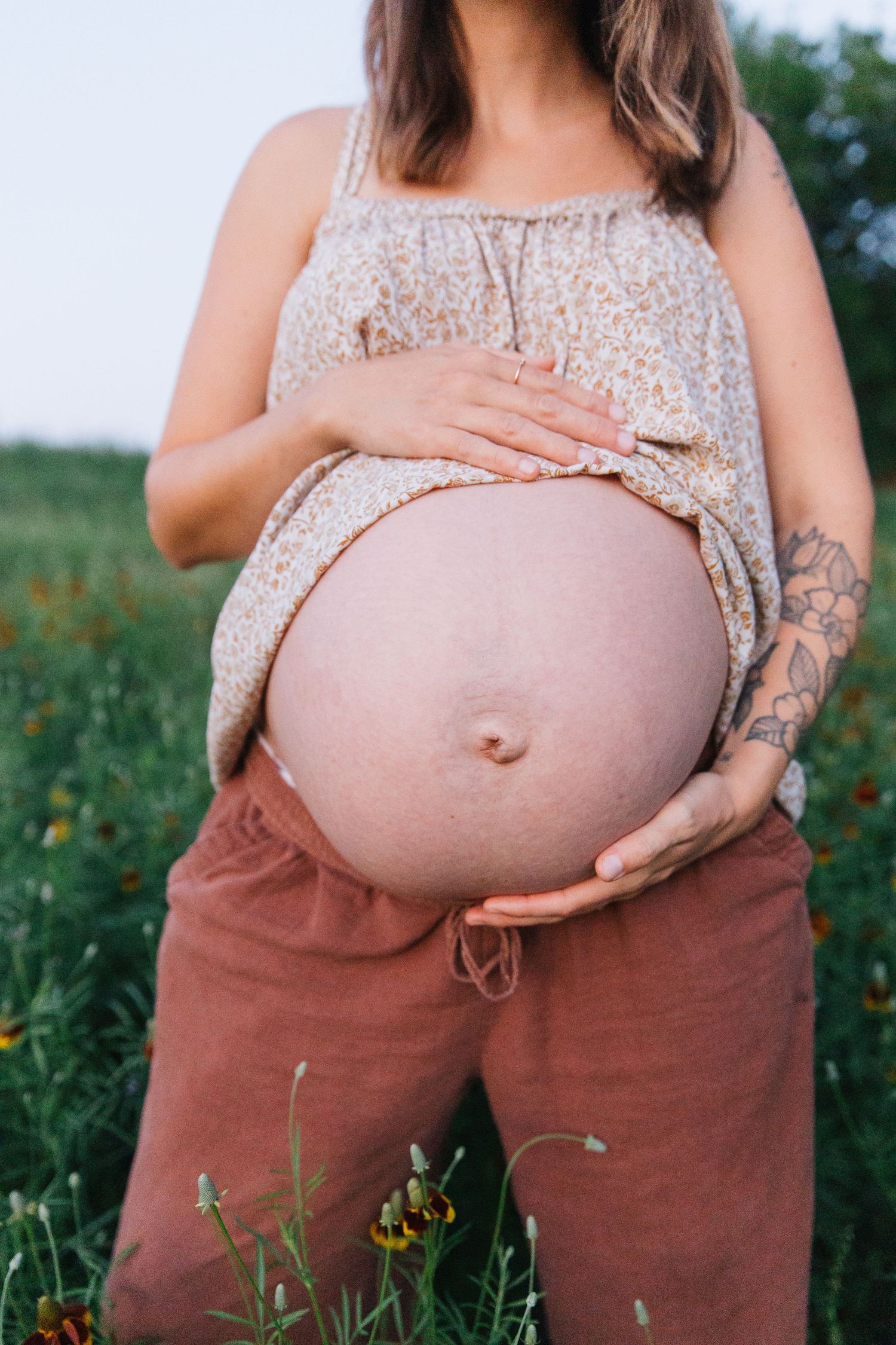 Pregnant woman in floral top and rust pants cradling bare baby bump in a green flower field.