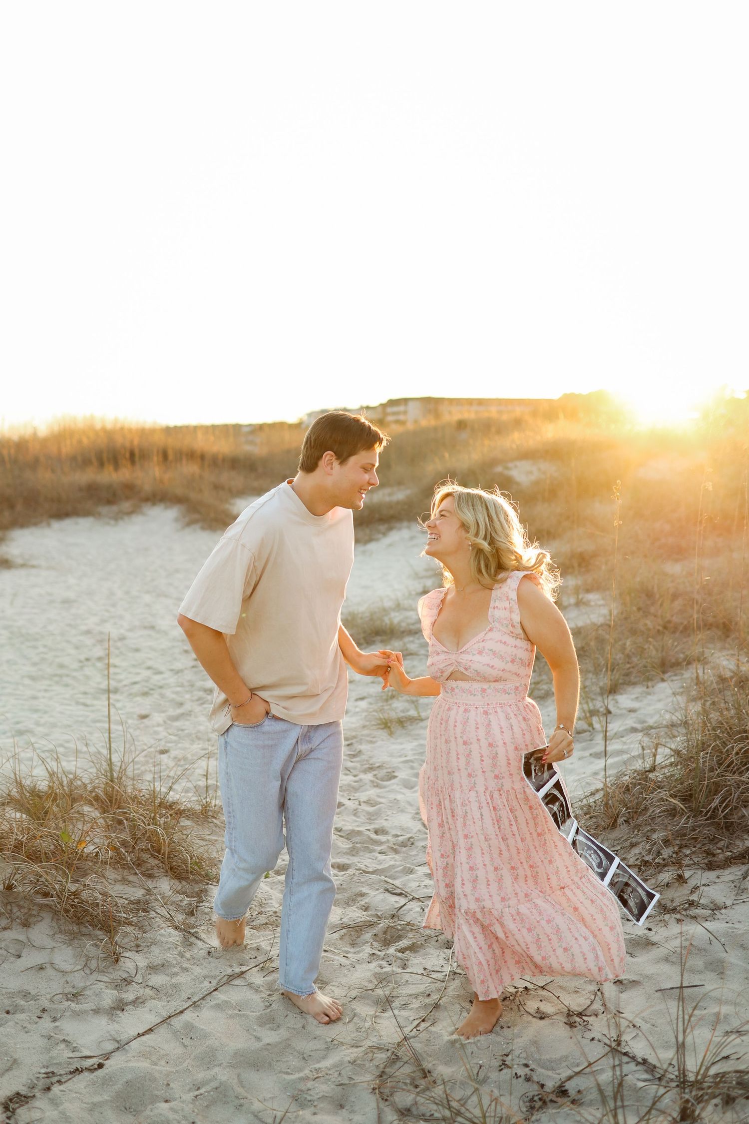 Hilton Head maternity photography with couple running through sand dunes holding ultrasound photos at Folly Field Beach