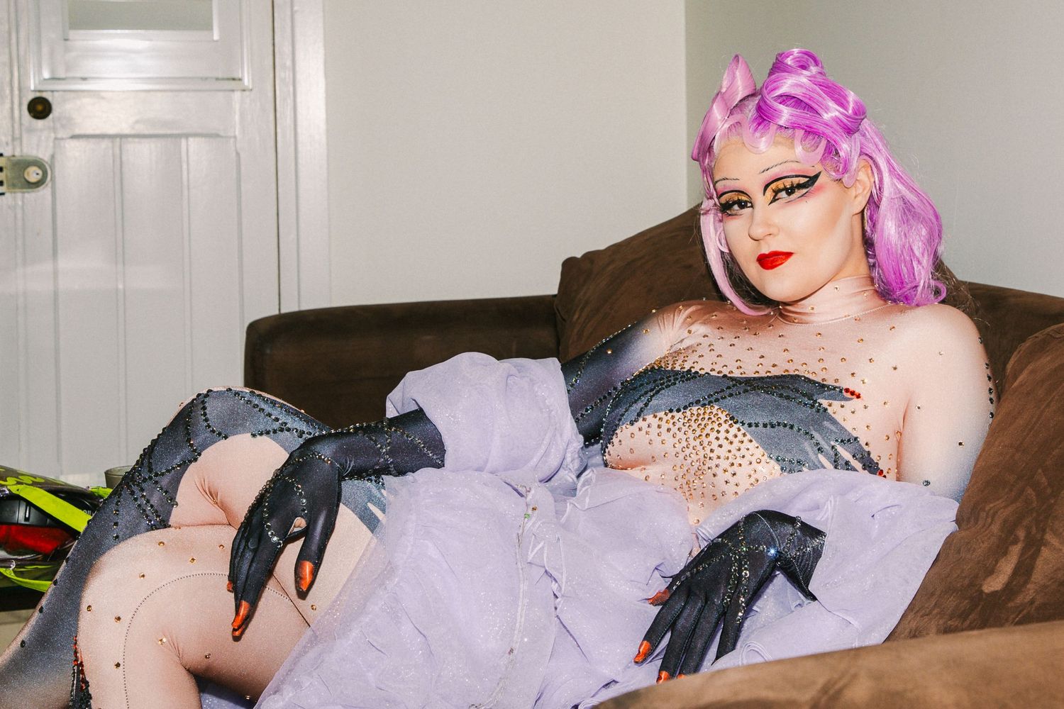 Drag performer in purple tulle dress and black gloves with pink hair posing on a couch at The Lass O'Gowrie Hotel.
