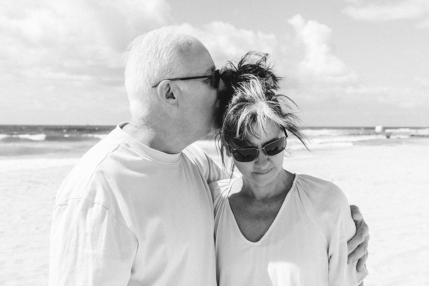 Elderly couple sharing a tender moment on a sunny beach, man kissing woman's head, both wearing white.