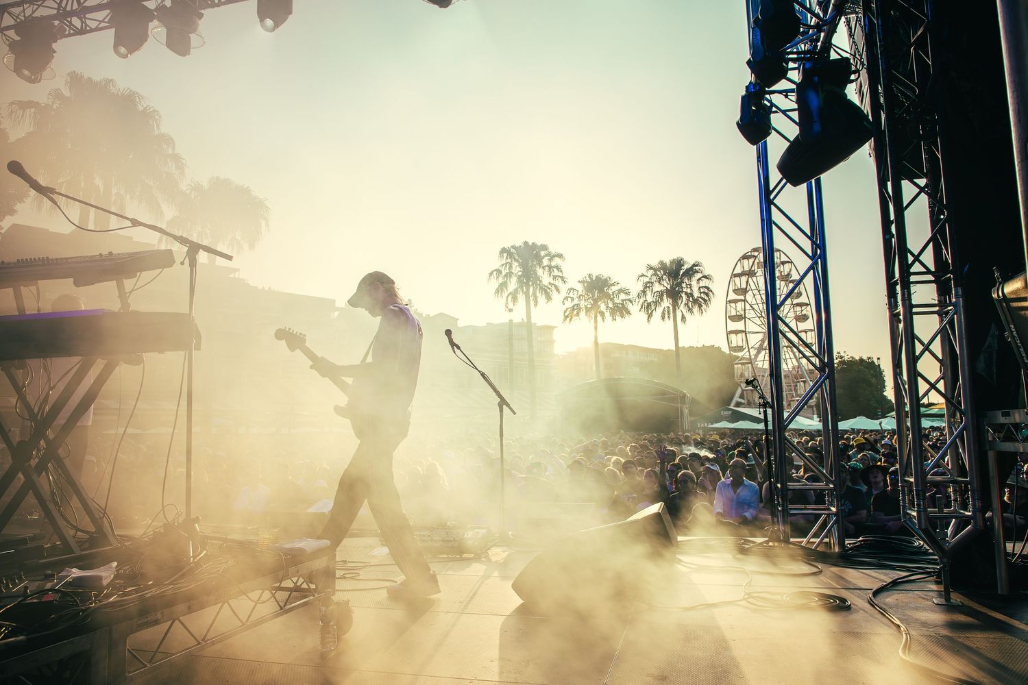 Silhouetted guitarist of Birds of Tokyo performing on a hazy outdoor Earp festival stage with palm trees in the background.