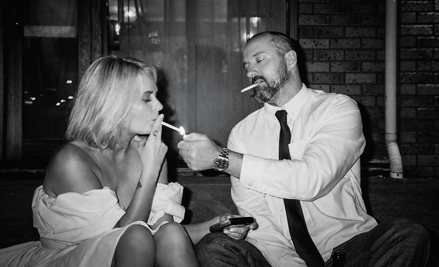 Black and white photo of bride and groom sitting together, man lighting womans cigarette at wedding reception.