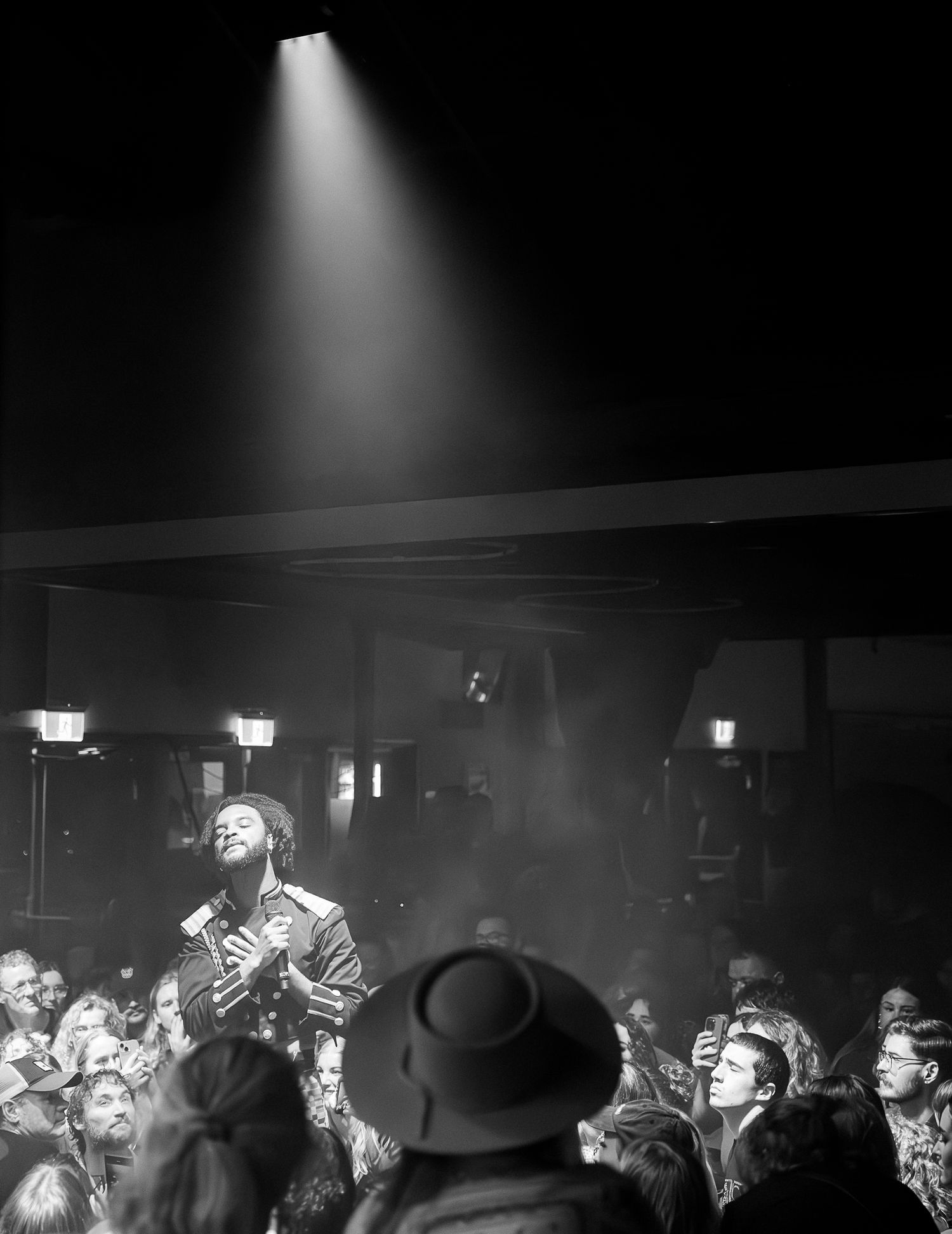 Black and white photo of a performer holding a mic under a spotlight beam in a crowded indoor venue.