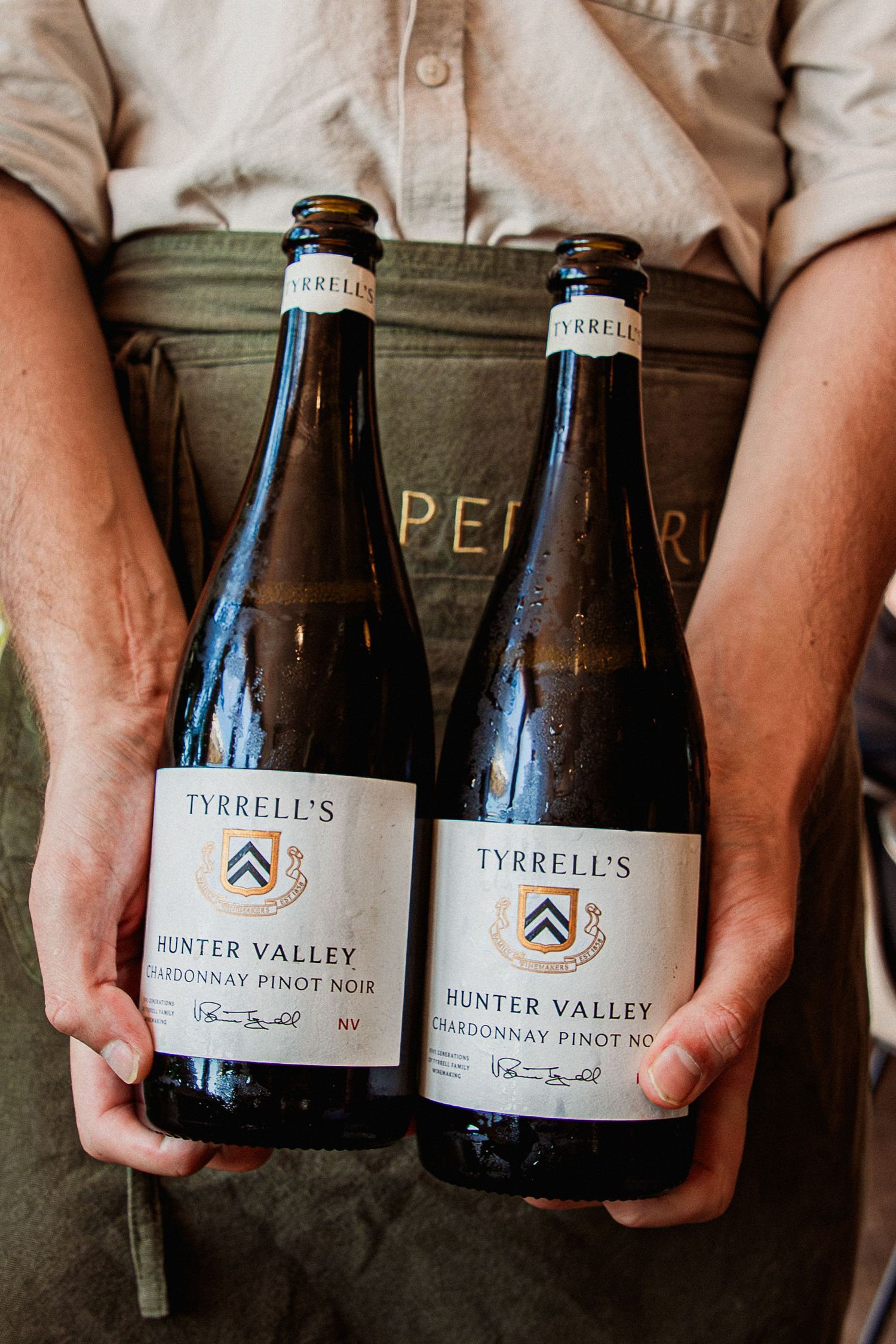 Person in a green apron holding two bottles of Tyrrell's Hunter Valley Chardonnay Pinot Noir wine at Peregrin Merewether.