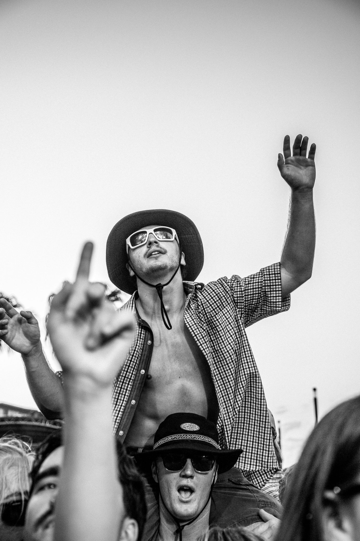 Black and white photo of shirtless man in hat sitting on shoulders in Earp festival crowd with hand raised.