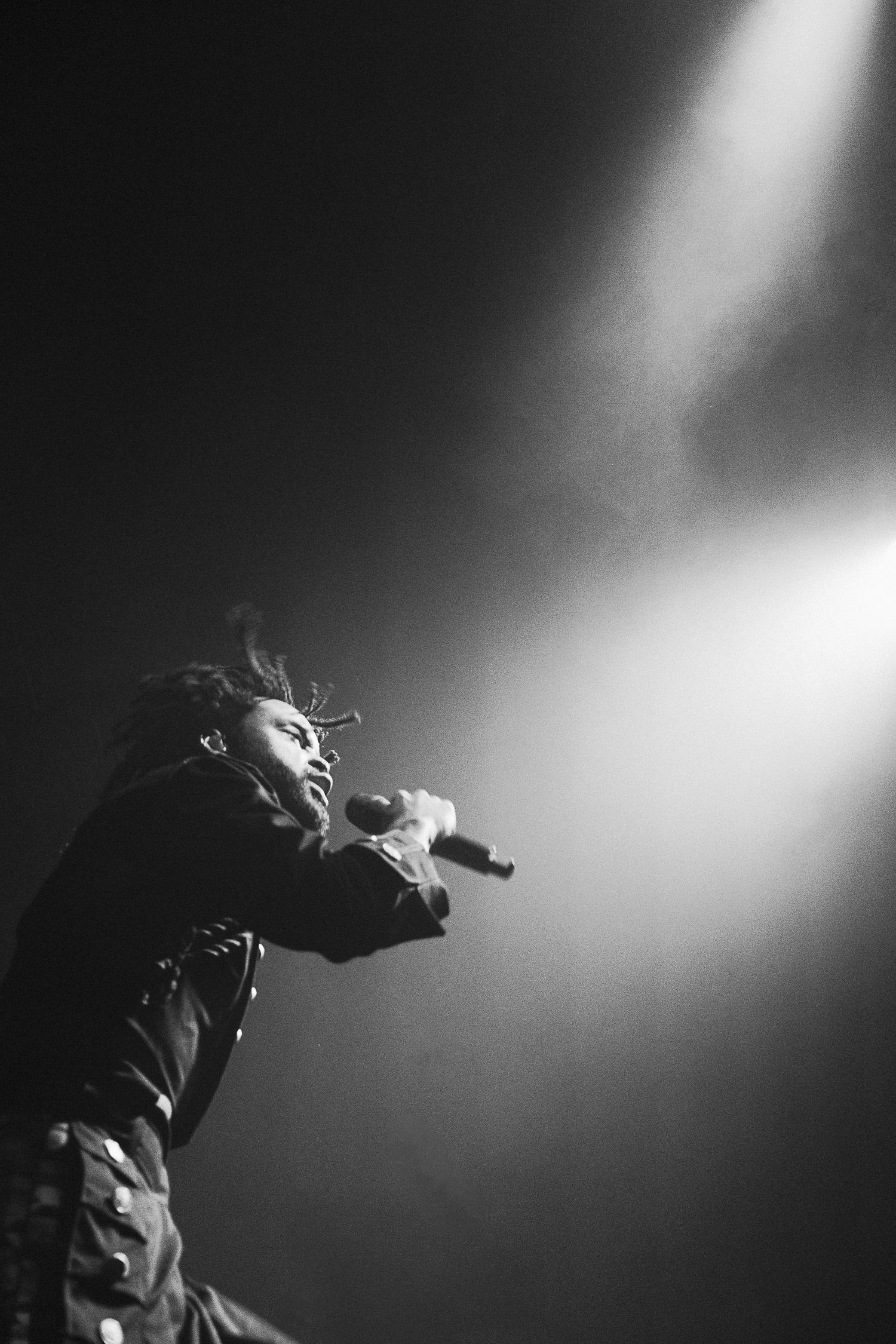 Black and white photo of a performer with dreadlocks singing into a microphone under dramatic stage lighting.