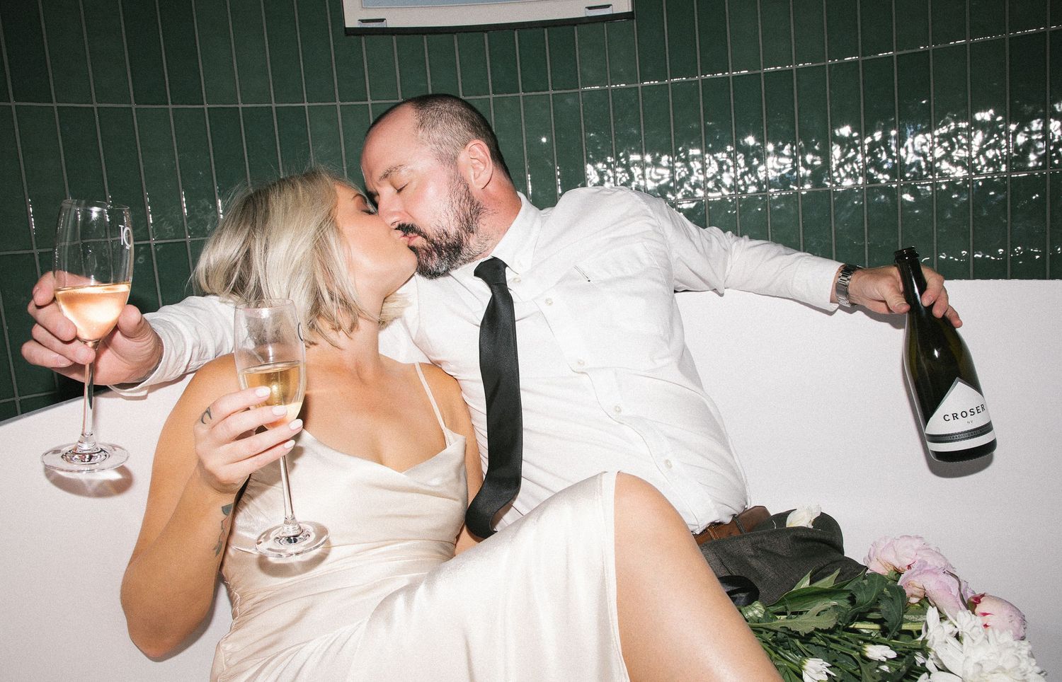 Newlyweds kissing in bathtub celebrating with champagne glasses and bottle in elegant wedding attire.