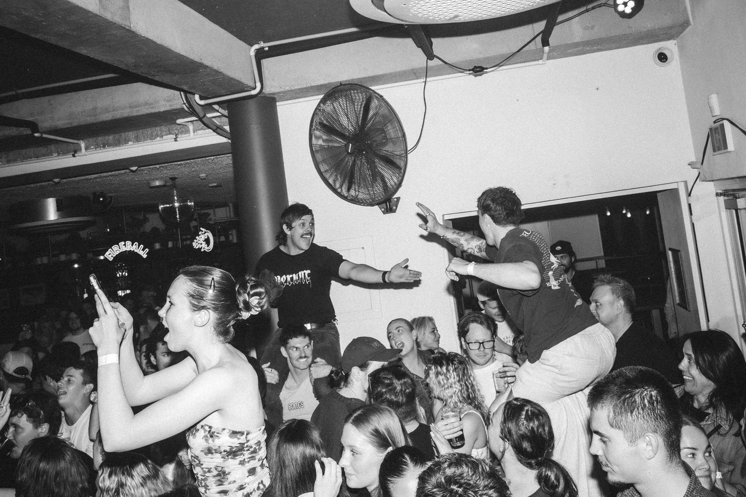 A energetic crowd surrounds a performer crowd surfing at an indoor black and white concert event at The Beach Hotel Merewether.