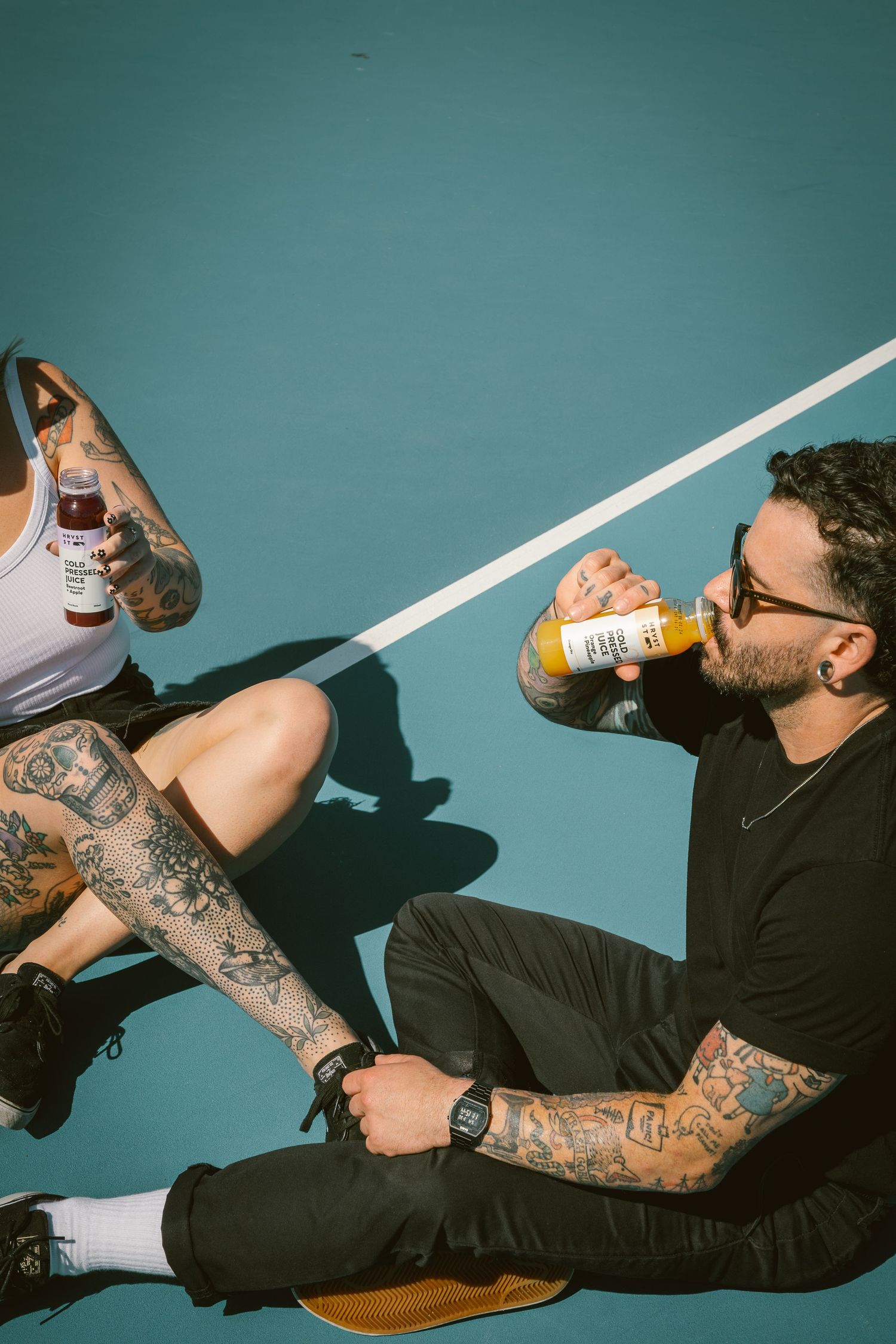 Two tattooed people relaxing on a teal tennis court drinking HRVST ST Cold Pressed Juice in sunny weather.
