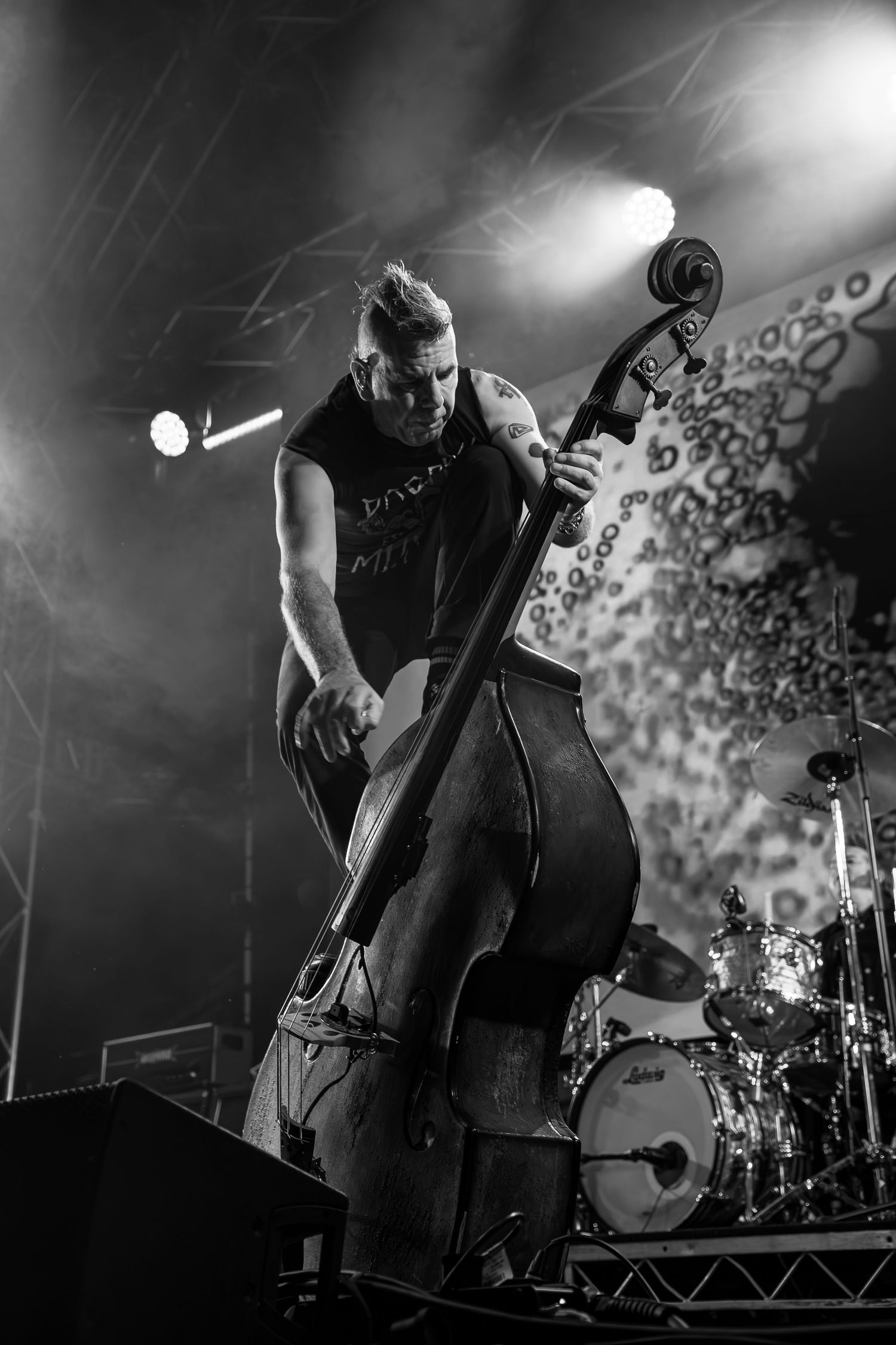 Black and white photo of the tattooed double bassist from The Living End energetically playing upright double bass on a concert stage.