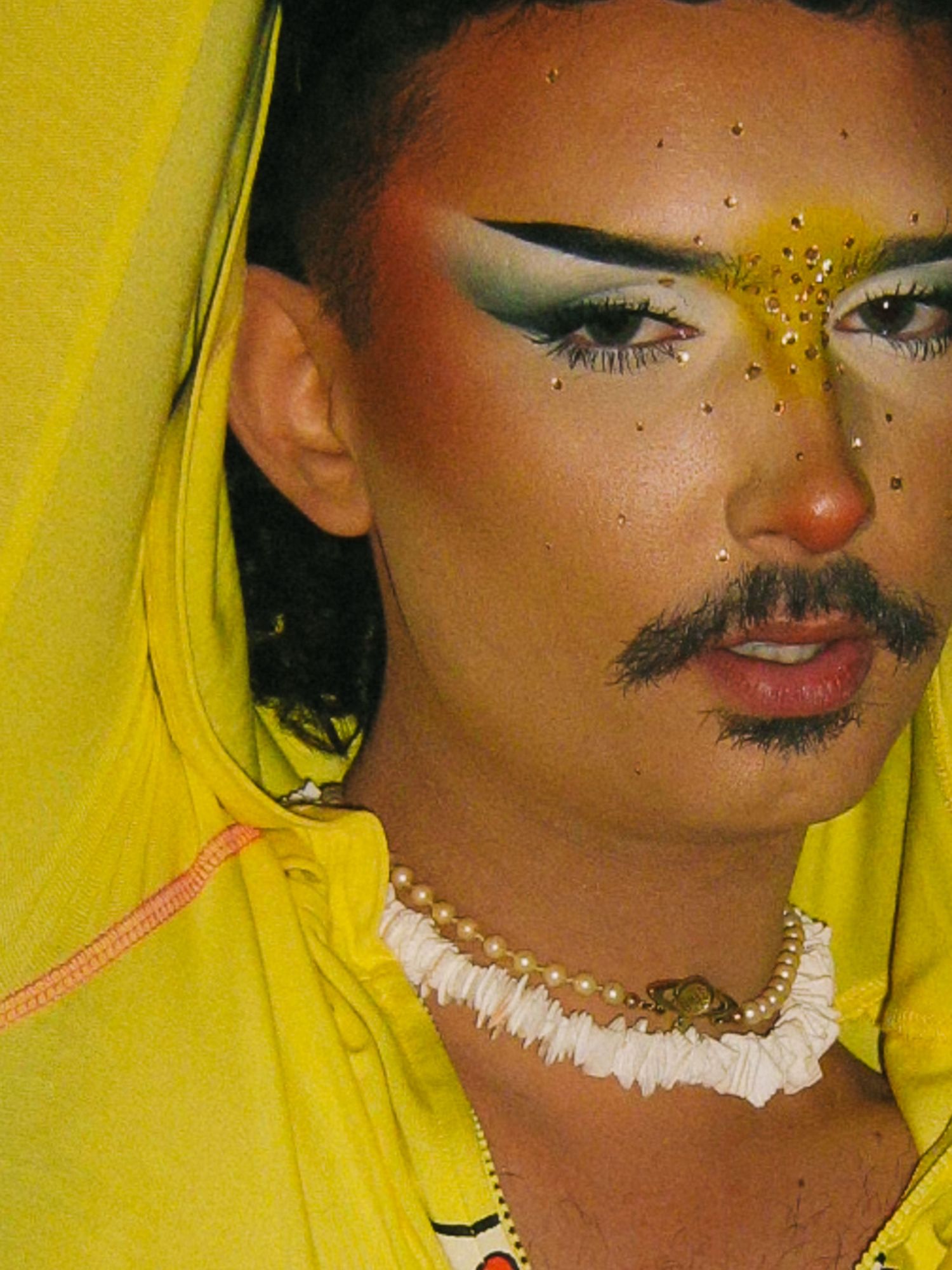 Close-up portrait of a person with dramatic eye makeup, mustache, gold glitter, and pearl necklace at The Lass O'Gowrie Hotel.