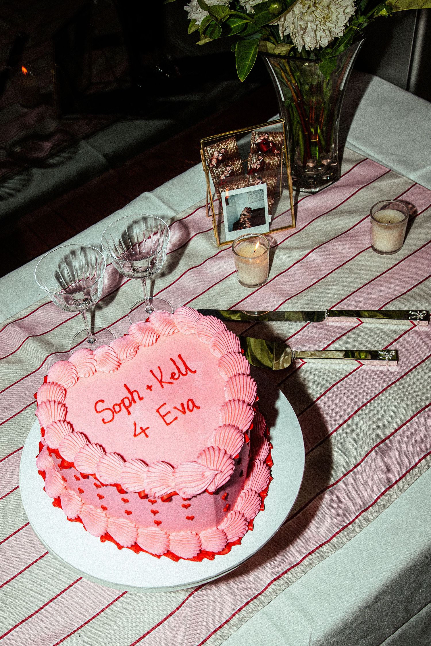 Pink heart-shaped wedding cake reading Soph and Kell 4 Eva on striped tablecloth with flowers and candles.