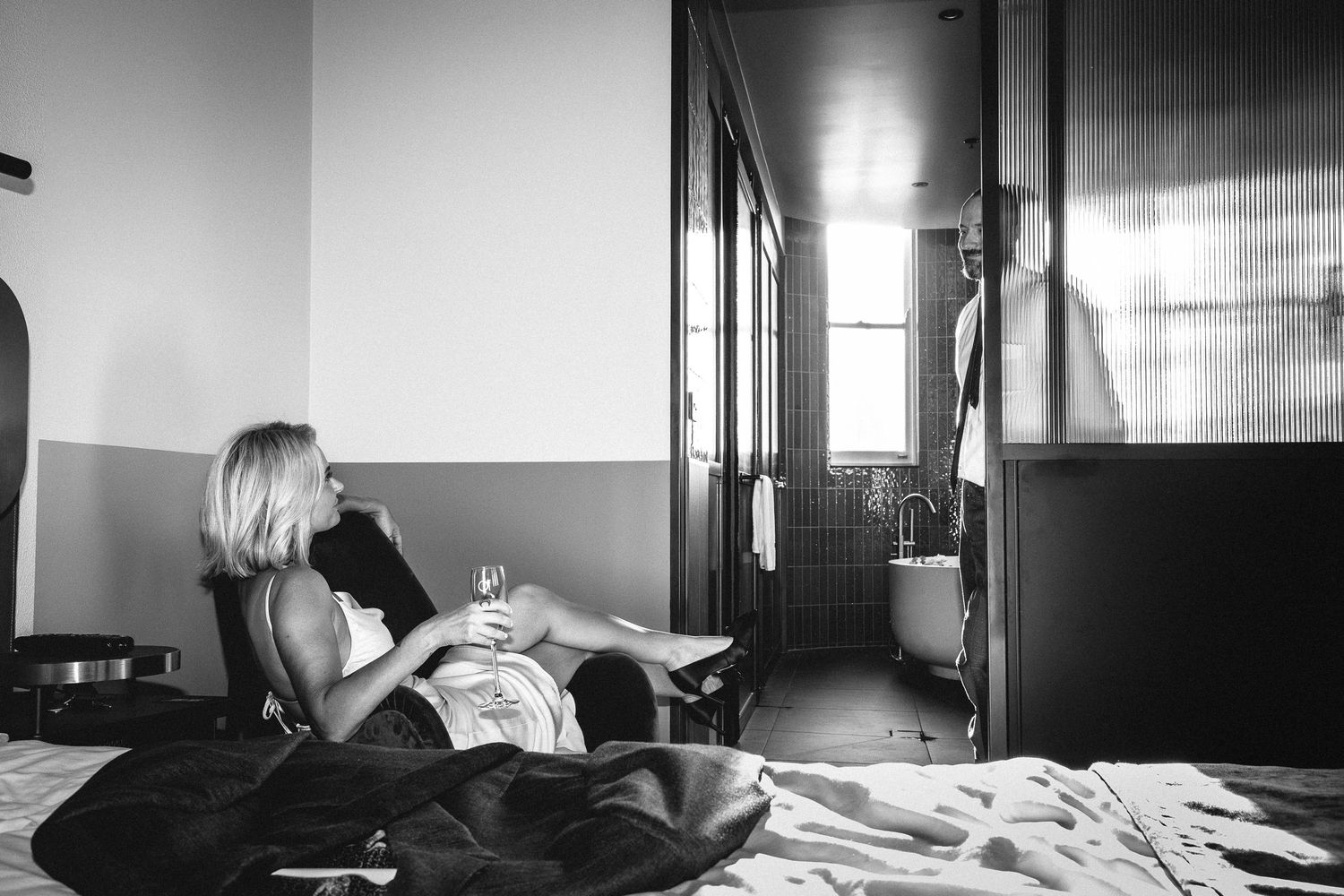 Bride in white dress relaxing on hotel bed with champagne glass before intimate elopement wedding.