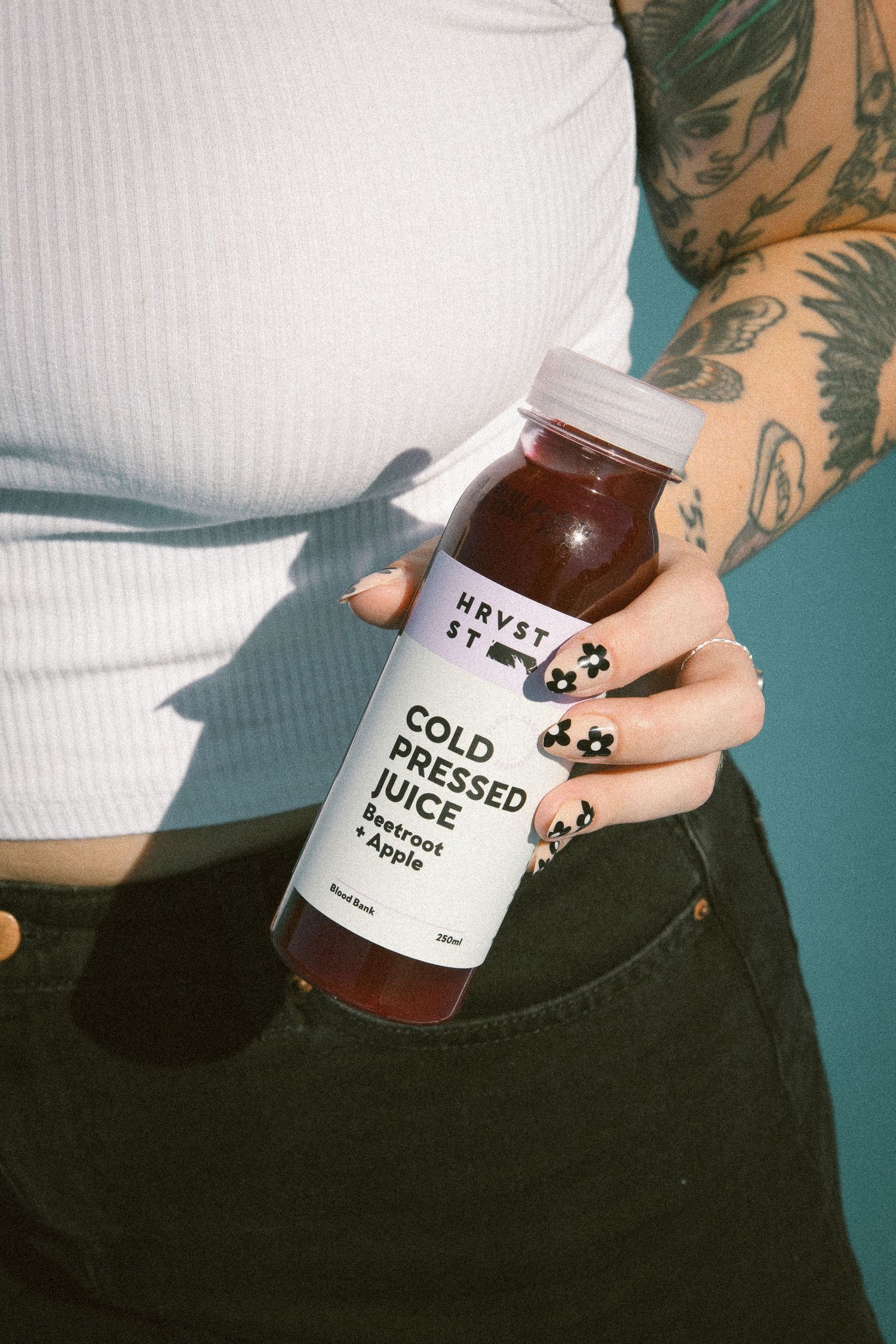 Tattooed woman in white crop top holding HRVST ST Cold Pressed Juice Beetroot and Apple bottle.