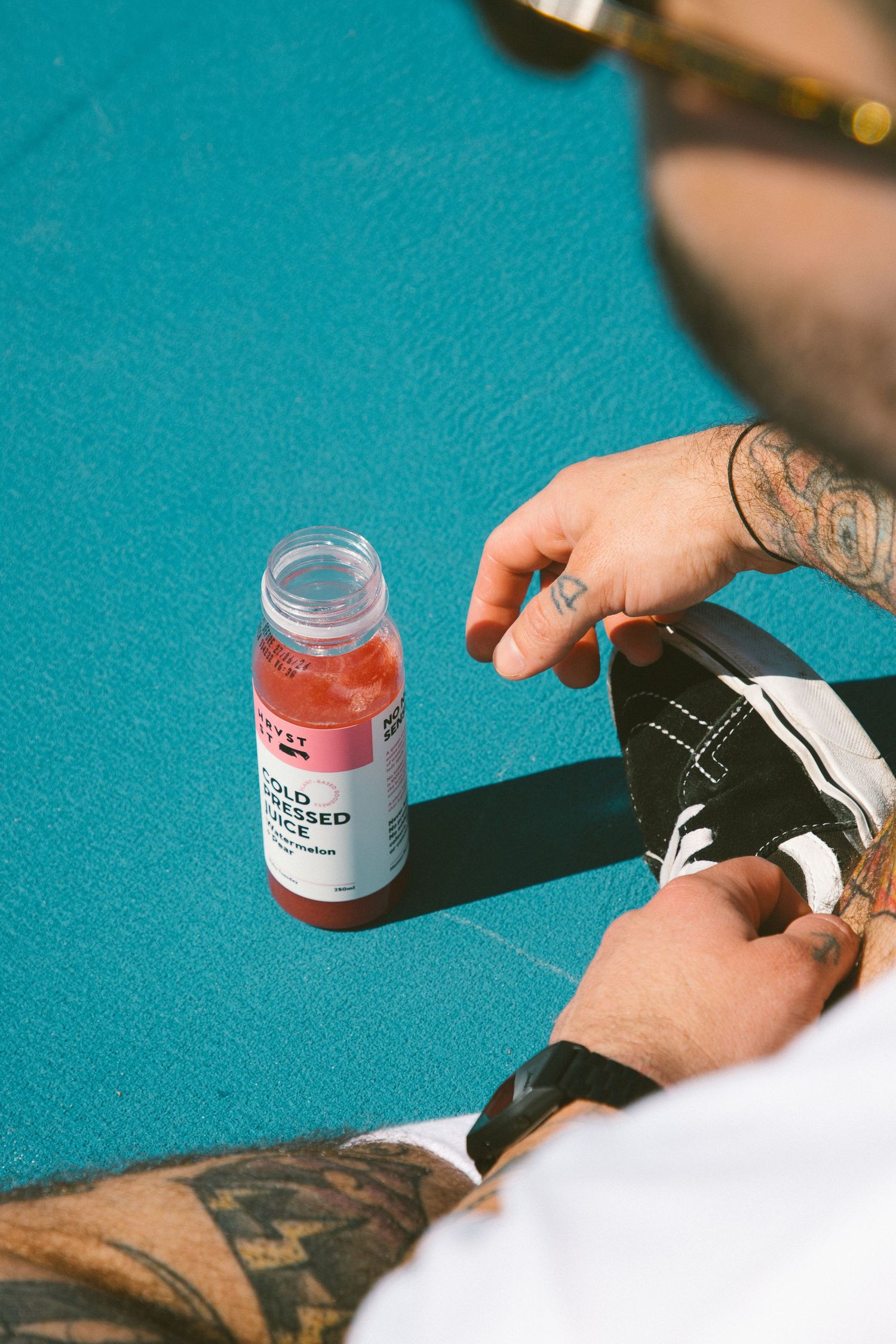 Cold pressed juice bottle on blue court surface with tattooed hand reaching nearby.