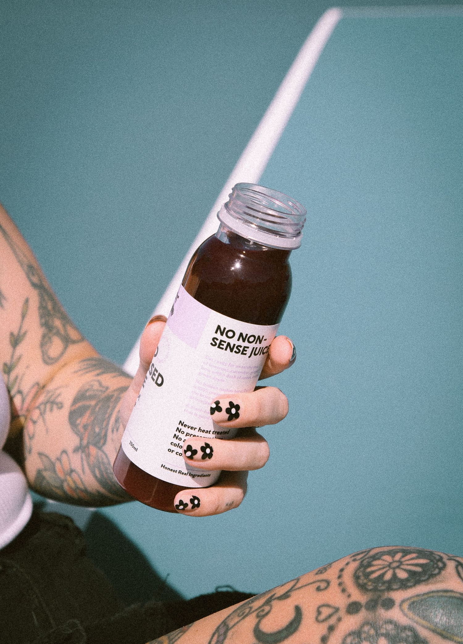 Tattooed hand with floral nails holding a No Non-Sense cold pressed juice bottle against teal wall.