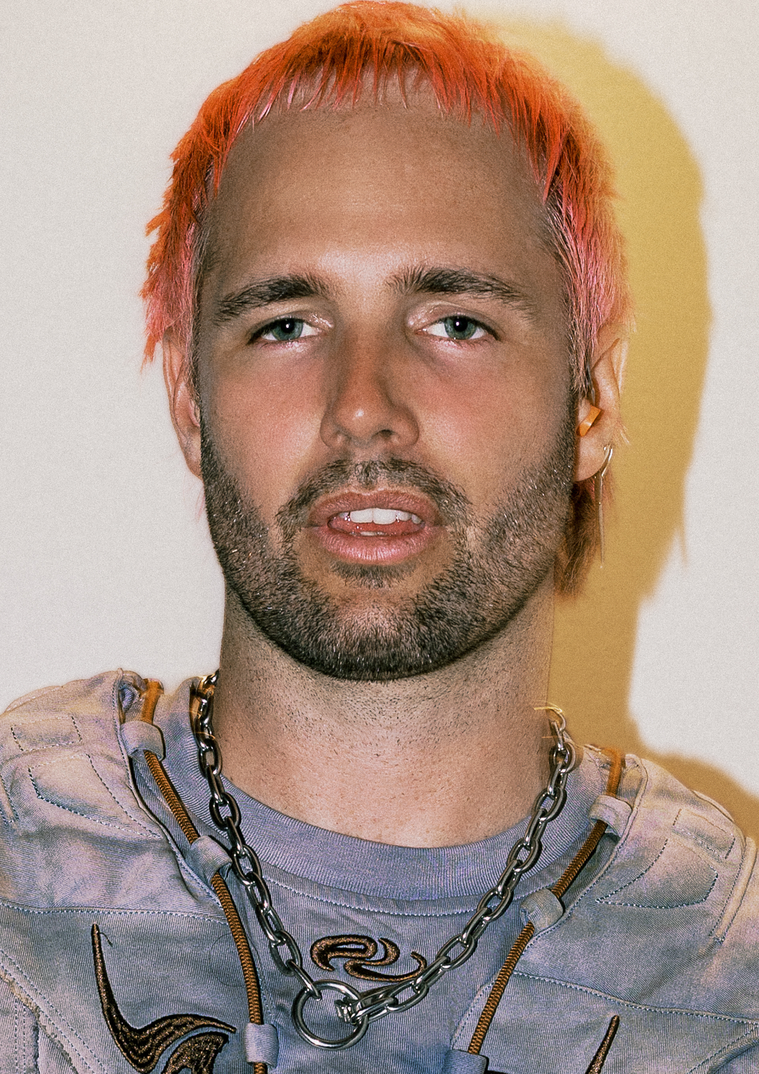 Young man with orange-dyed hair, stubble beard, wearing chain necklace and graphic tee against cream background.