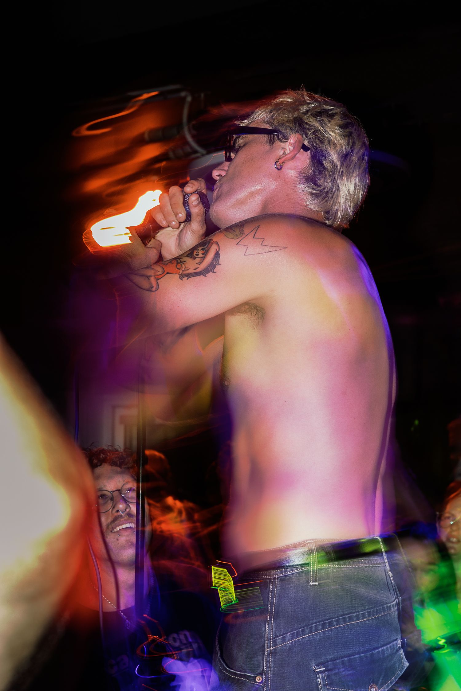 Shirtless tattooed performer sings into microphone under colorful stage lights at a nighttime concert event.