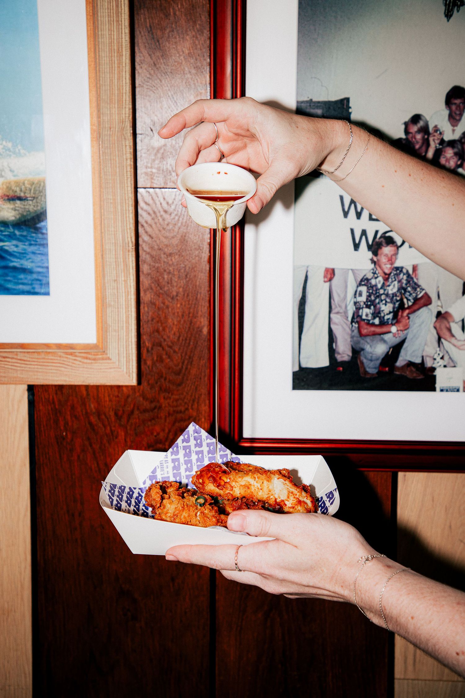 Hands holding a paper tray of fried chicken pieces and dipping sauce near framed photos on a wooden wall at Larrie's Takeaway Merewether.