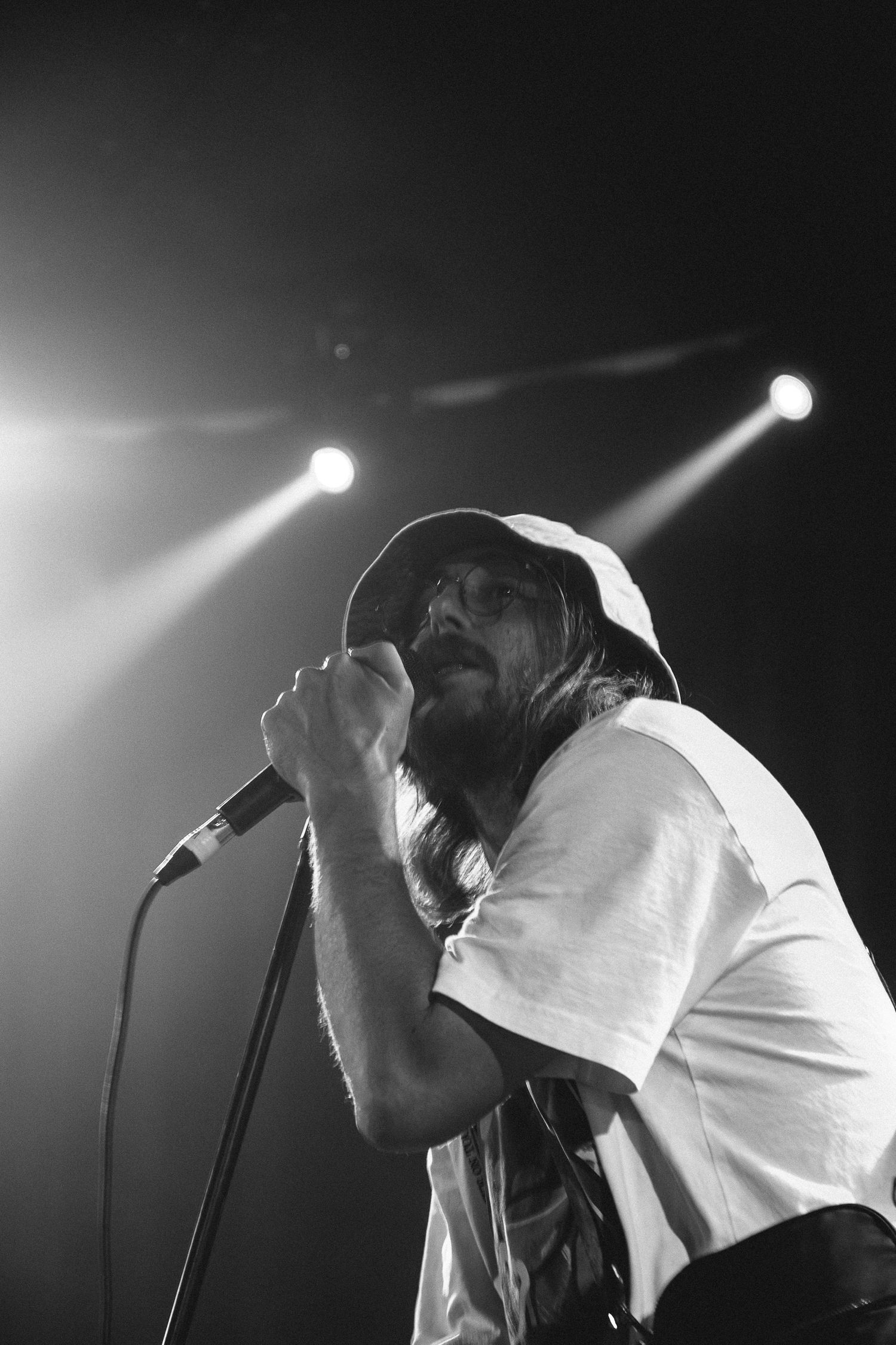 Black and white photo of bearded male singer performing on stage with mic, wearing bucket hat and white t-shirt under dramatic spotlights.