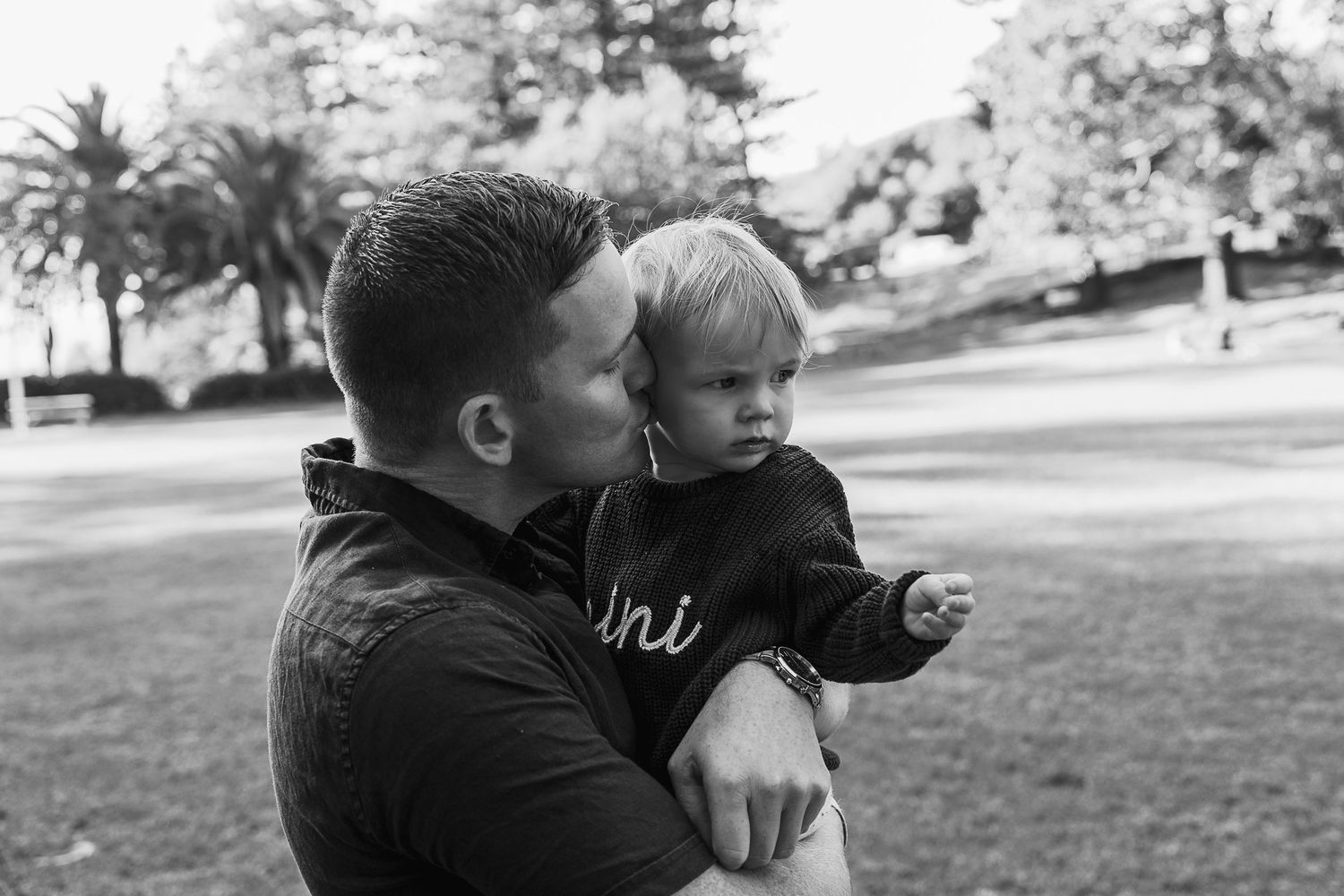 Black and white photo of a father kissing his toddler child outdoors in a park setting.