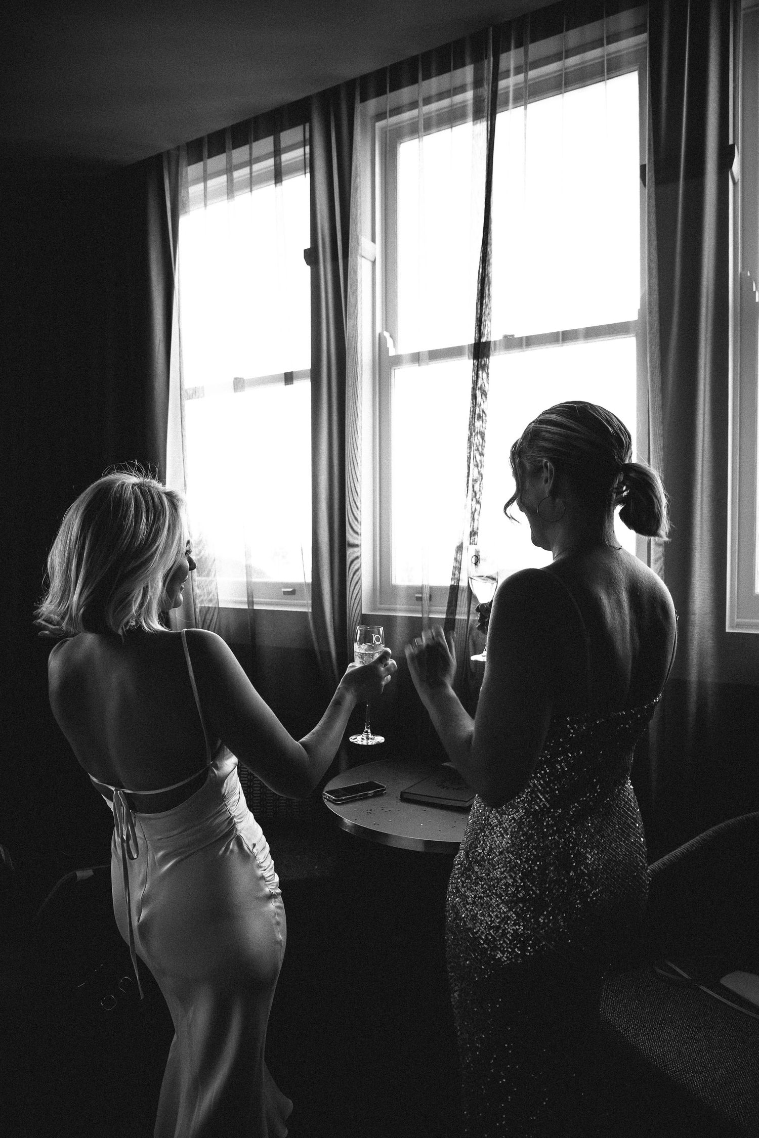 Black and white silhouette of bride and bridesmaid toasting champagne glasses by sunlit hotel window.