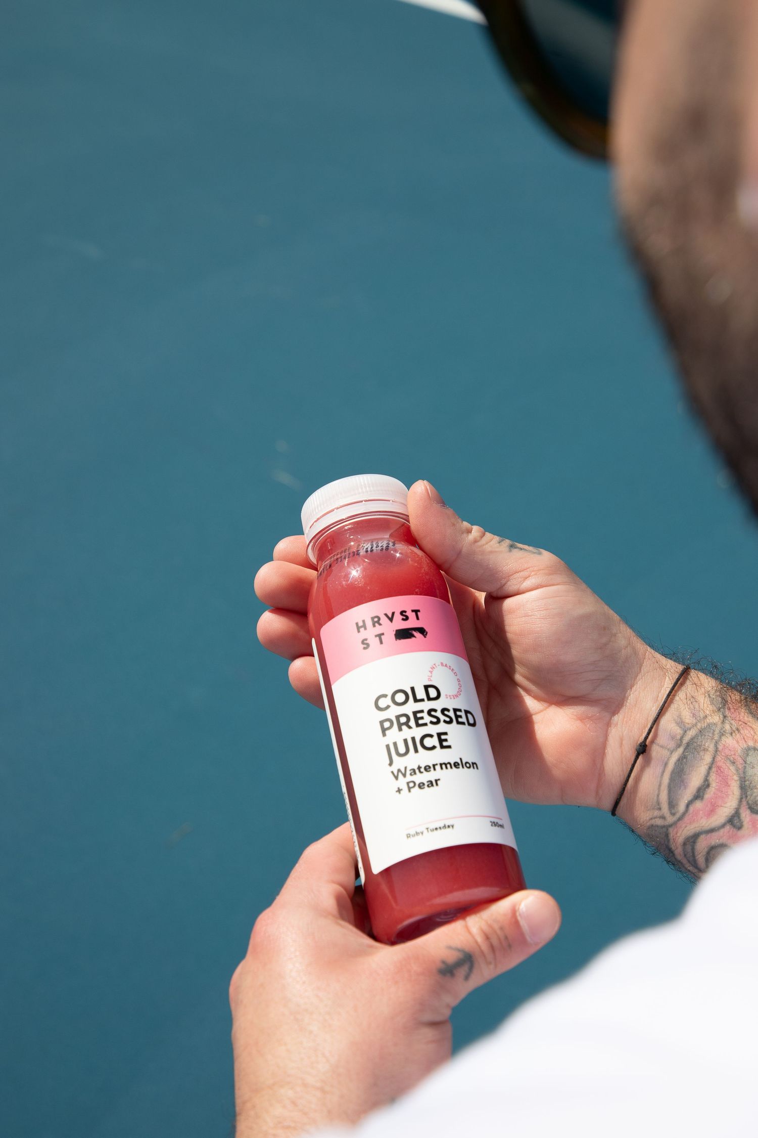 Close-up of tattooed hand holding Harvst St cold pressed watermelon and pear juice bottle.