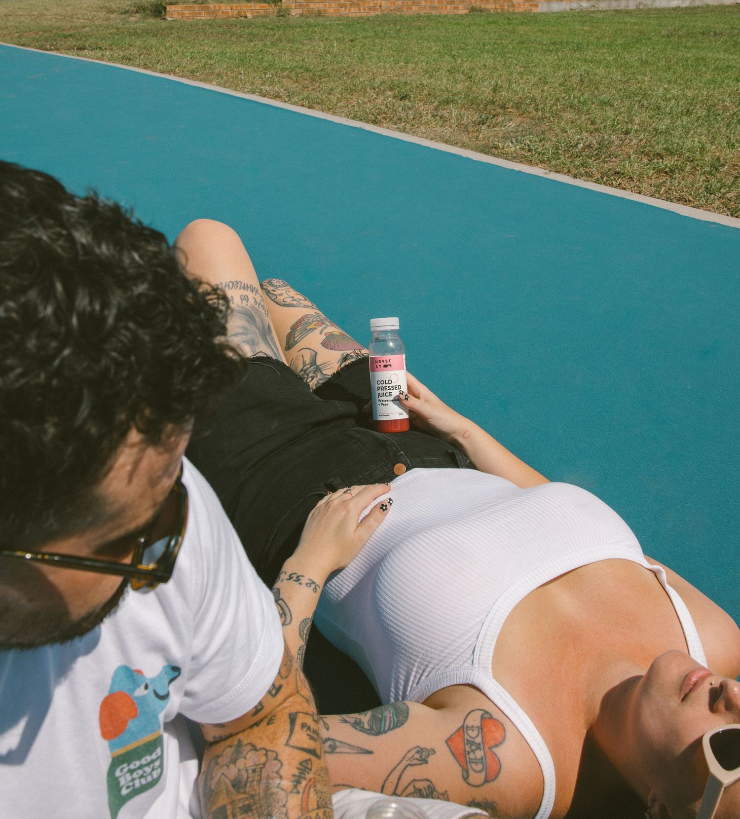Tattooed couple relaxing on a blue outdoor court, woman in white bodysuit holding a cold pressed juice bottle.
